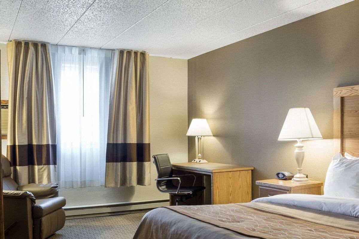 A hotel room with a bed, a desk, a chair, a nightstand, and curtains.