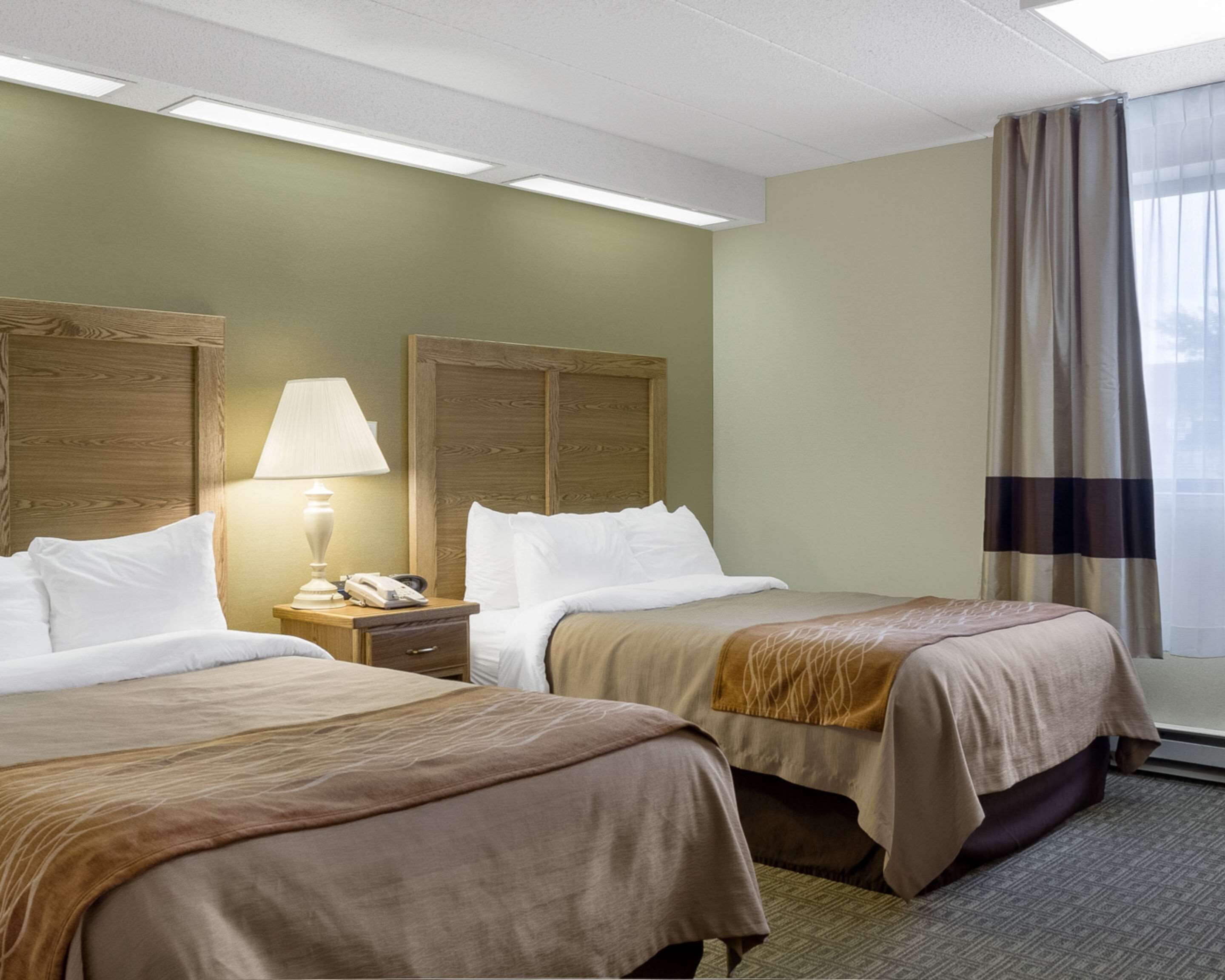 A hotel room with two beds, a nightstand with a lamp and phone, and a window with curtains.