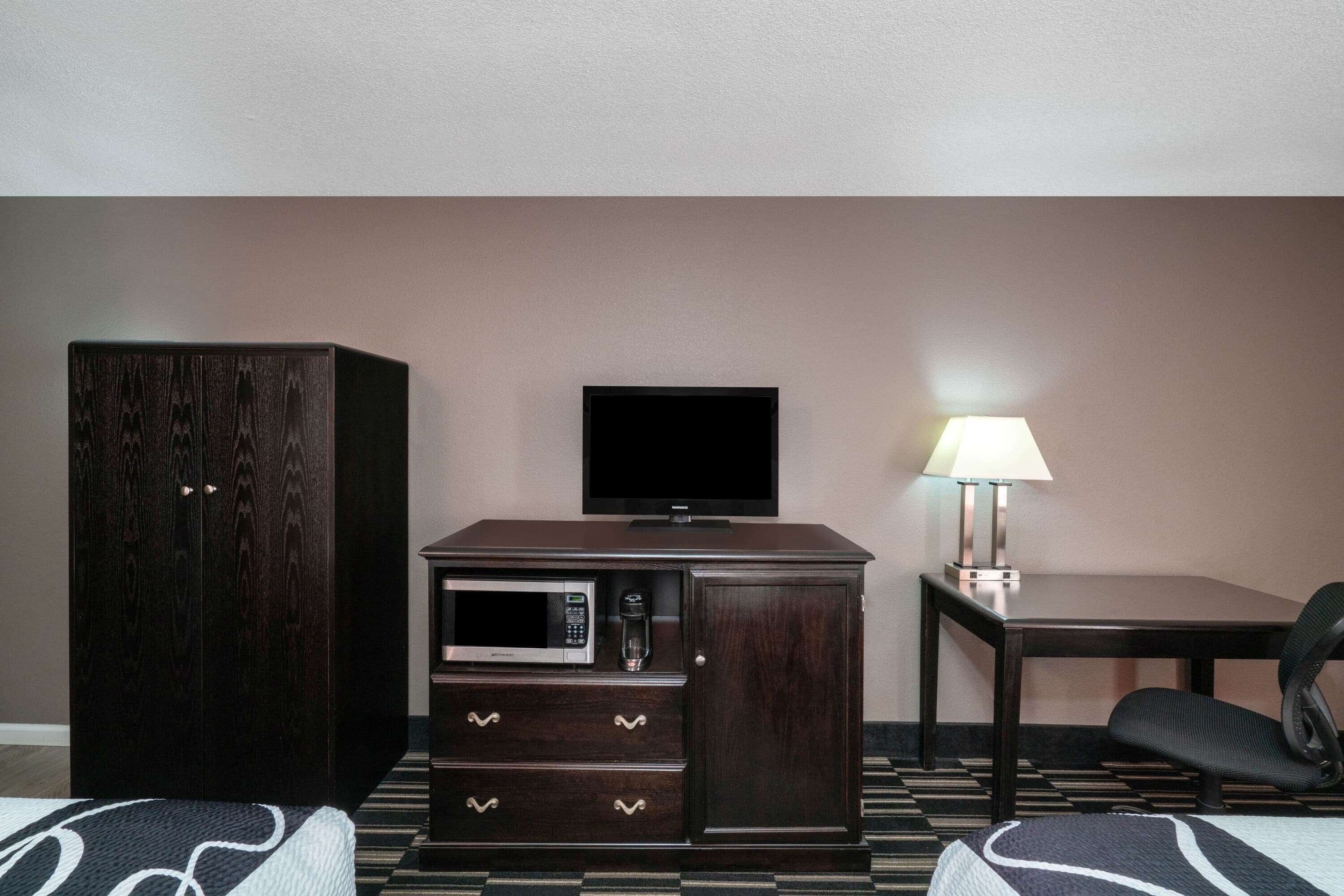 Camera d'albergo con un mobile scuro in legno, una TV a schermo piatto, un forno a microonde, un tavolo con una lampada e una sedia.