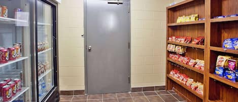 A small room with a refrigerator stocked with beverages, a door, and shelves with snack items.