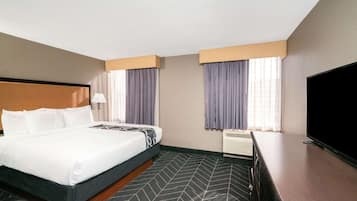 A hotel room with a bed, a desk, a flat-screen TV, and two windows with curtains.
