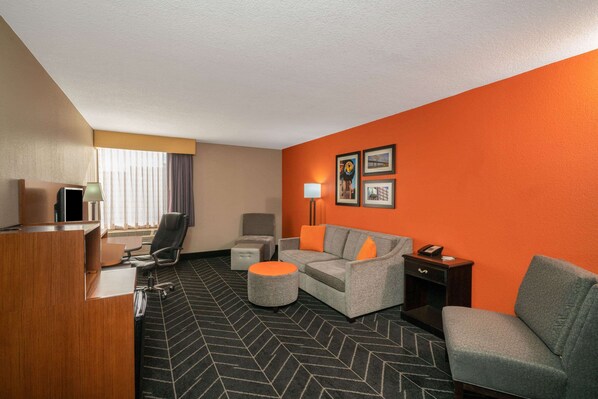 Habitación de hotel con un mostrador de recepción, un sofá gris con cojines naranjas, un sillón gris, un pequeño reposapiés, una lámpara de pie y cuadros en una pared naranja.