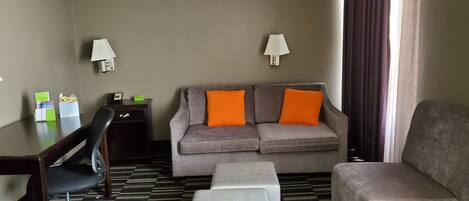 Ein Hotelzimmer mit grauem Sofa, einem Schreibtischstuhl, einem kleinen Tisch, einer Lampe und einem Fenster mit Vorhängen.