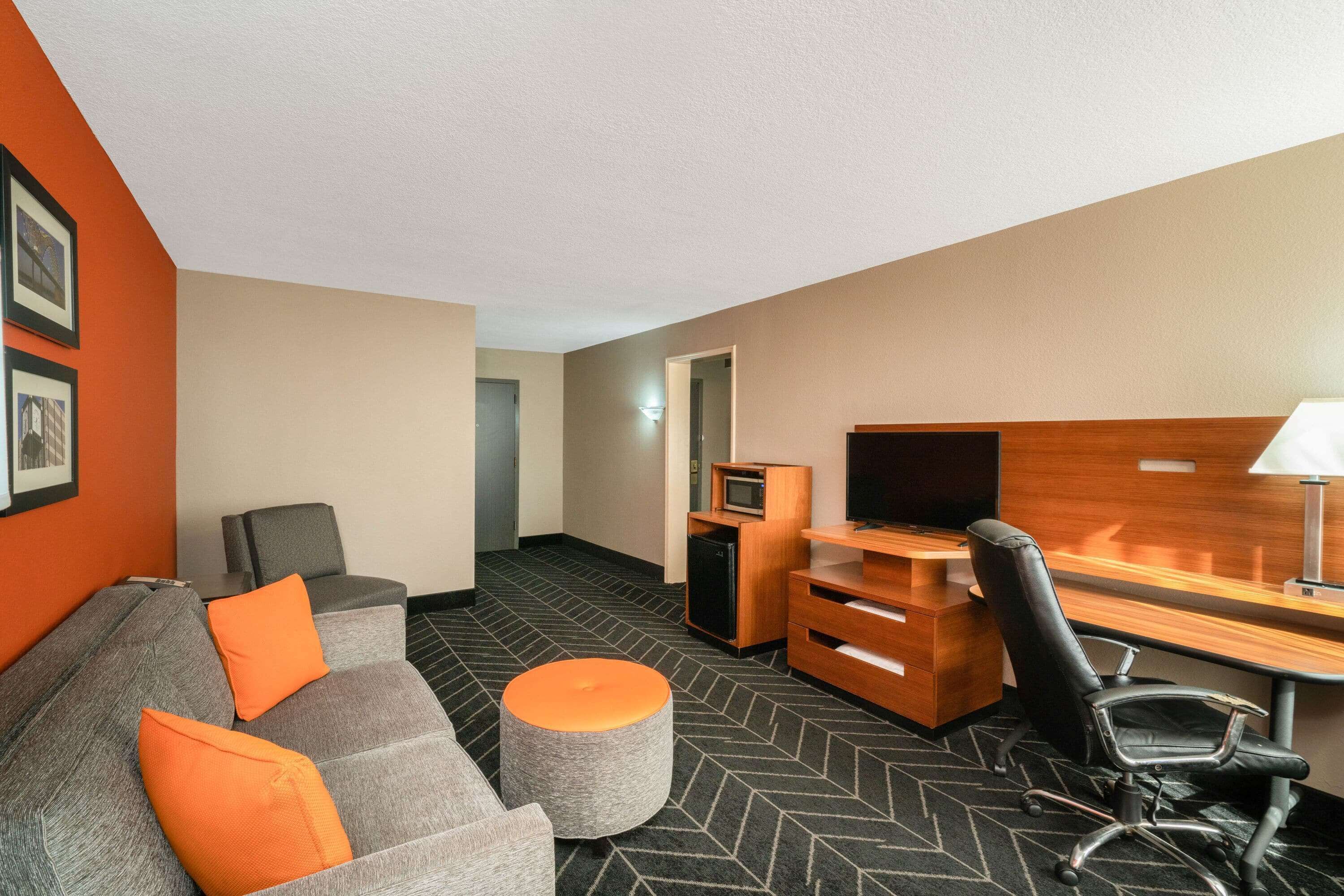 A hotel room with a grey sofa, orange cushions, a wooden desk, a black office chair, a small ottoman, a flat-screen TV, and a microwave.
