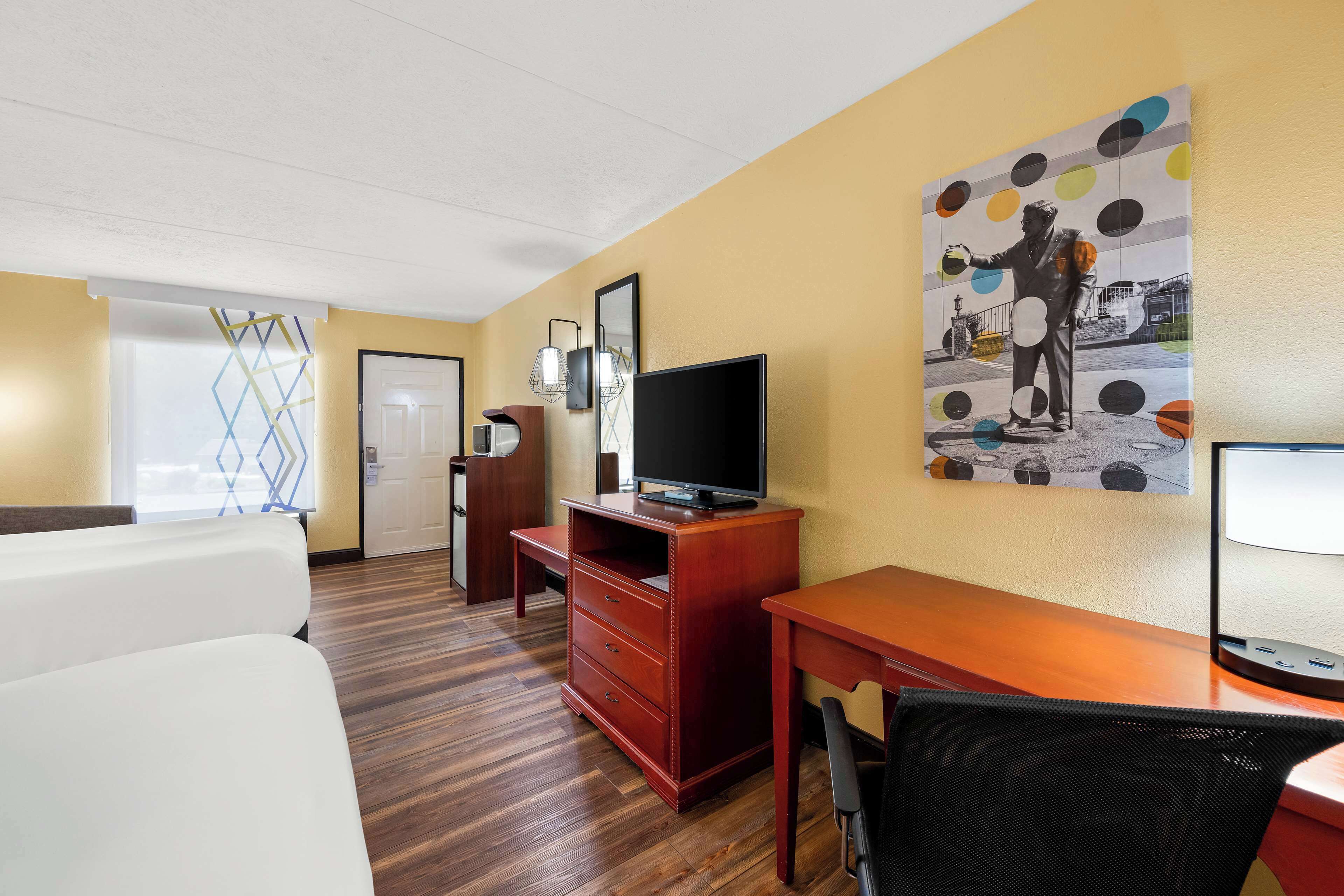 Une chambre d’hôtel avec un plancher en bois, un téléviseur posé sur un meuble en bois, un bureau avec une chaise et un tableau accroché au mur.