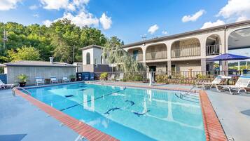 A swimming pool with lounge chairs and umbrellas, surrounded by a building with a covered patio and a view of trees.