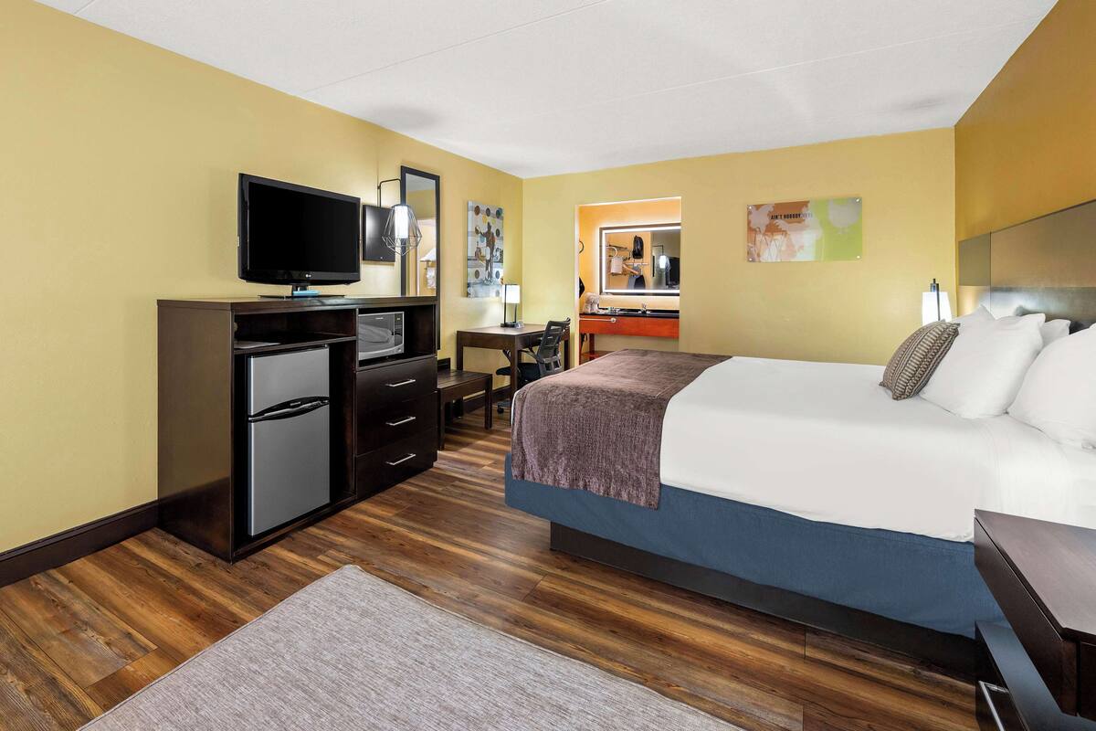 A hotel room with a bed, a desk with a chair, a television, a mini-fridge, and a bathroom with a mirror and sink.