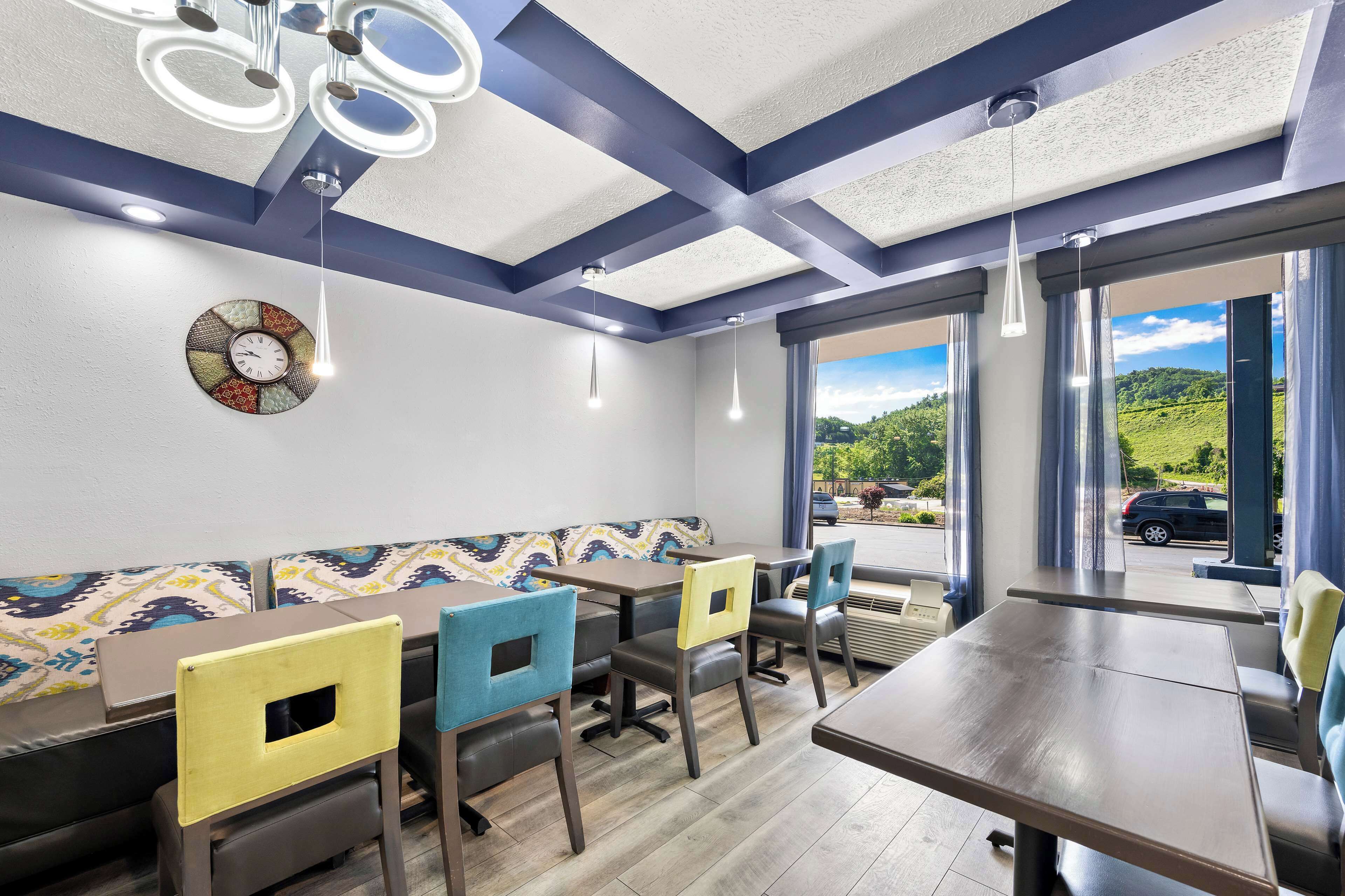 A restaurant with a modern interior, featuring a large dining table, multiple chairs, a patterned wall clock, and a view of the outdoors.