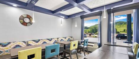 A restaurant with a modern interior, featuring a large dining table, multiple chairs, a patterned wall clock, and a view of the outdoors.