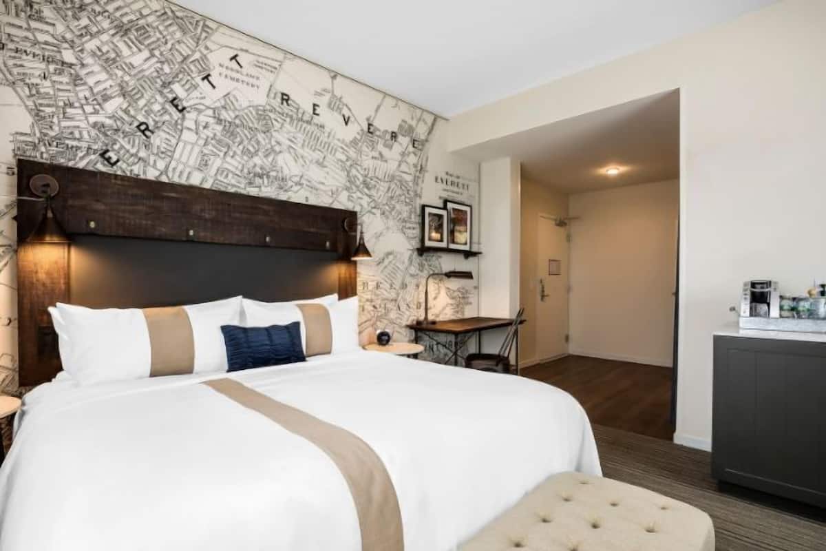 Ein Hotelzimmer mit einem großen Bett, einem Kopfteil, einem kleinen Tisch, einer Bank und einer Küchenzeile.