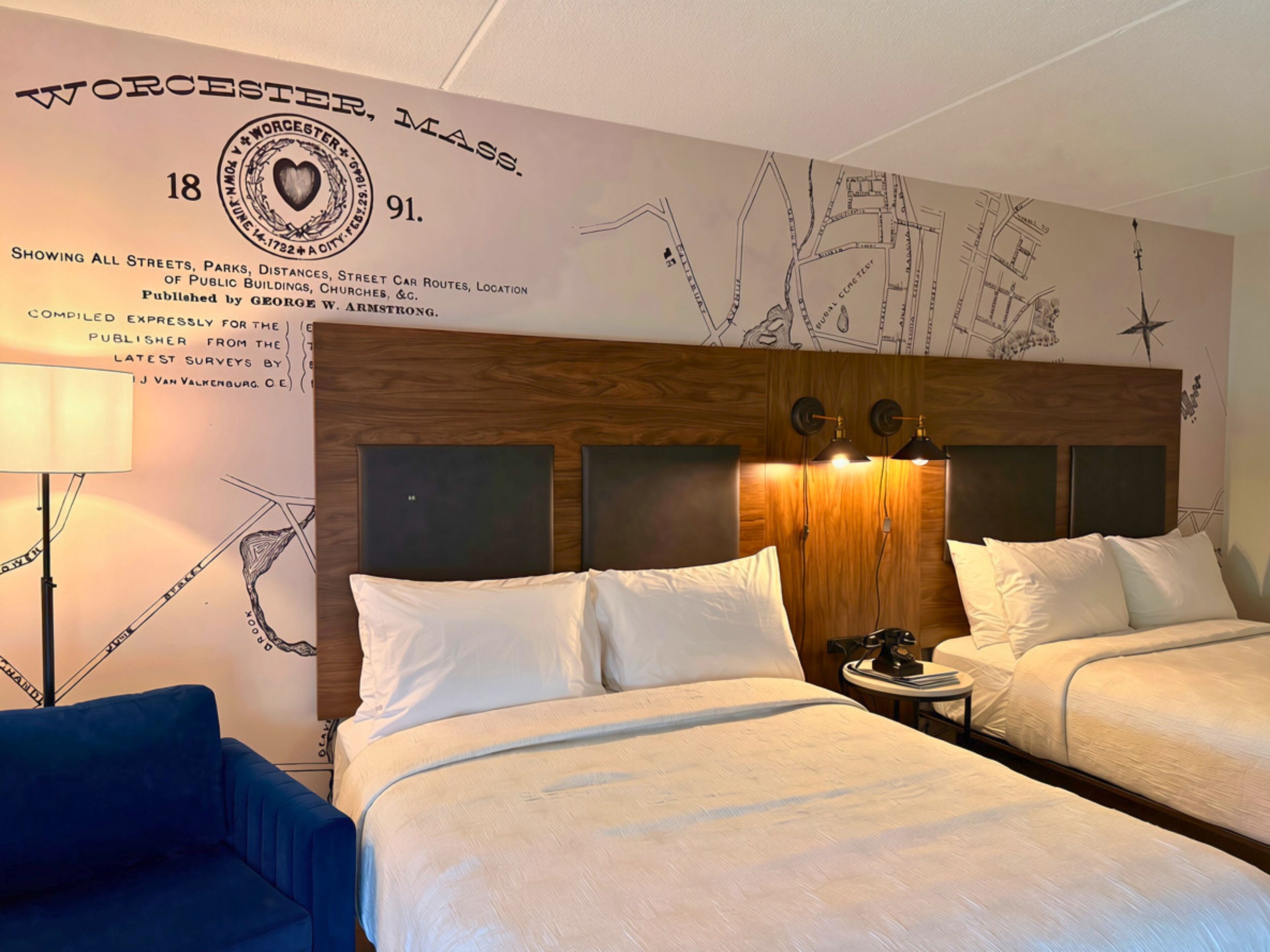 Habitación de hotel con dos camas, cabecero de madera, un mapa en la pared y una lámpara.