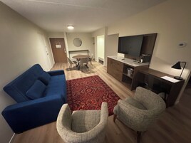 Una sala de estar con un sofá azul, dos sillones, una mesa de comedor con sillas, un televisor y un escritorio pequeño con una lámpara.