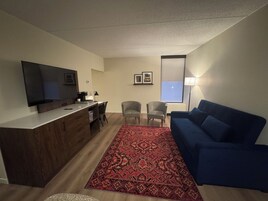 Una sala de estar moderna con un televisor de pantalla plana, un sofá azul, un tapete estampado y una zona de comedor con sillas y una cafetera.