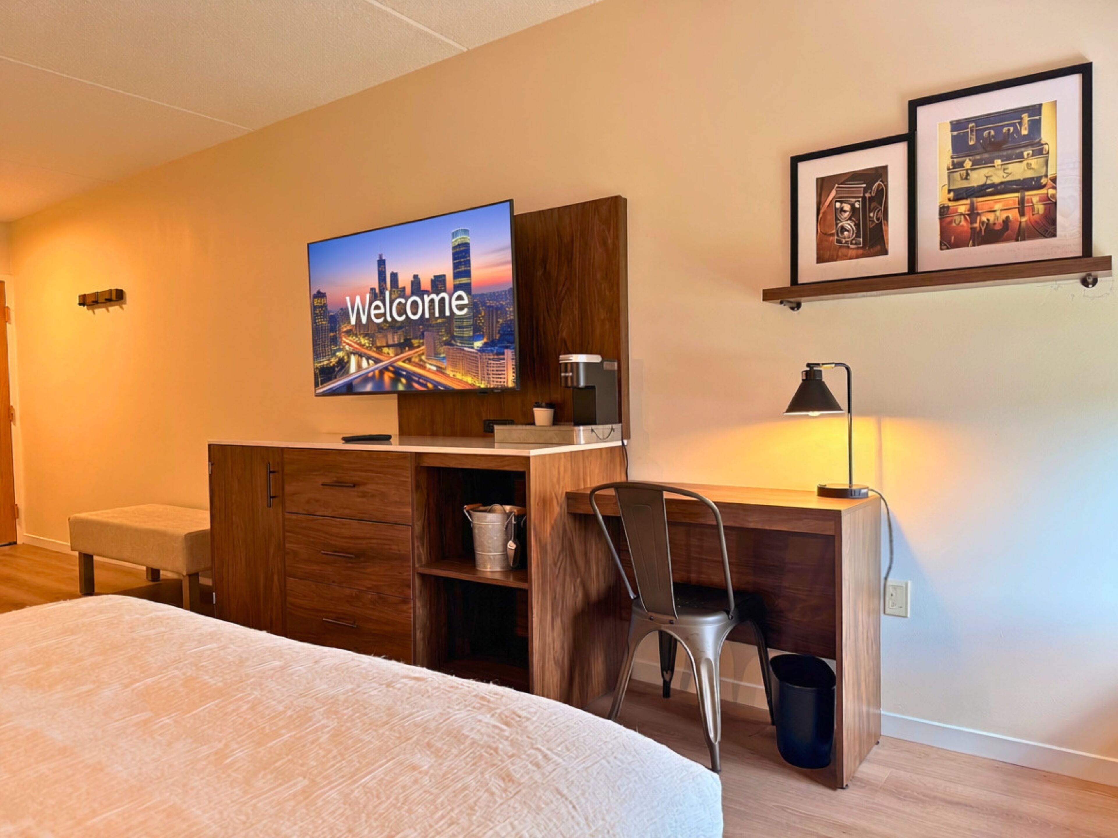 Una habitación de hotel con una cama, un escritorio con una silla, un televisor mostrando 'Welcome' y una estantería con fotografías enmarcadas.
