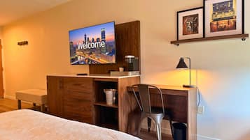 Una habitación de hotel con una cama, un escritorio con una silla, un televisor mostrando 'Welcome' y una estantería con fotografías enmarcadas.