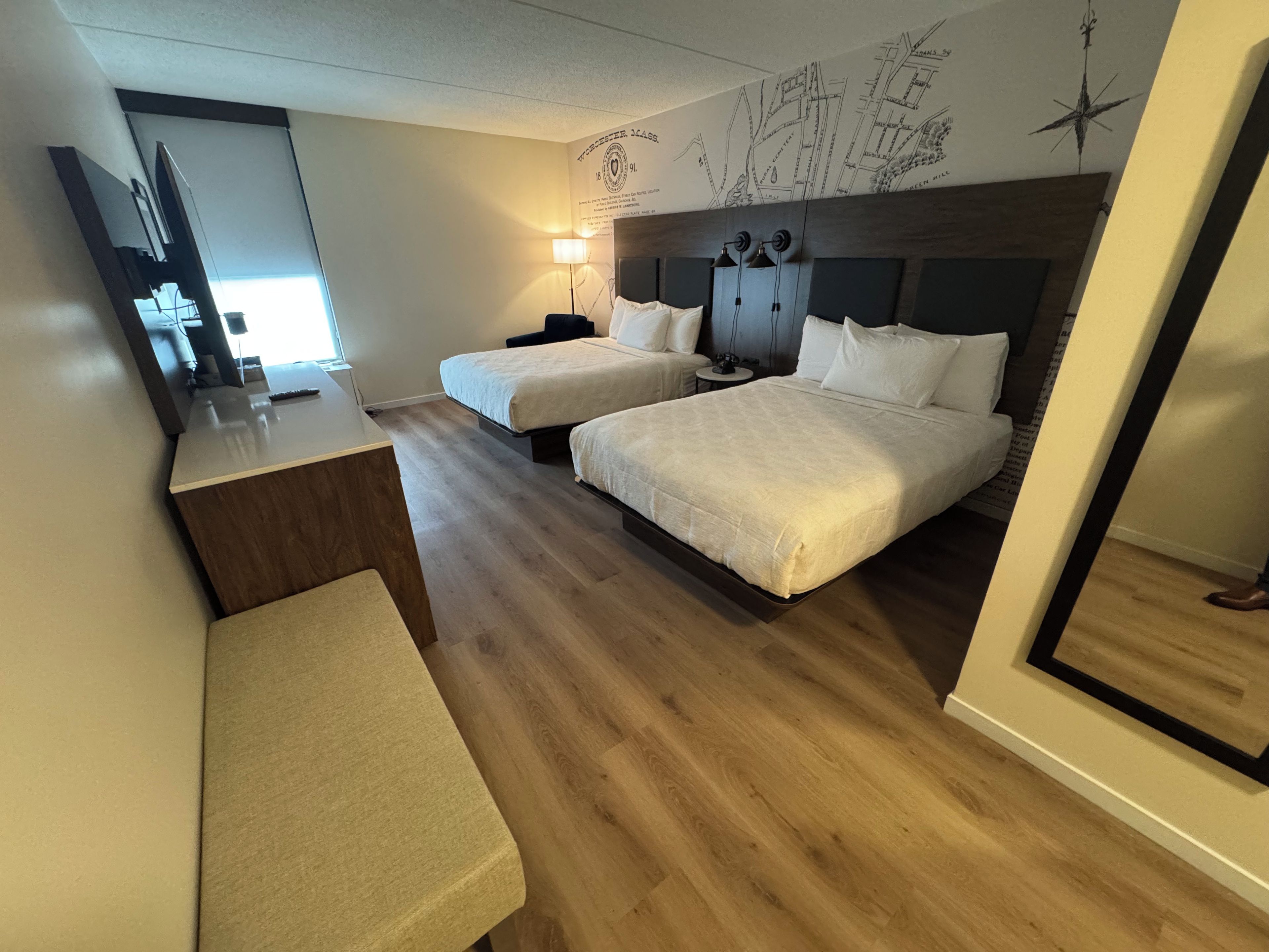 Quarto de hotel com duas camas, uma escrivaninha, um banco e um mapa de parede.