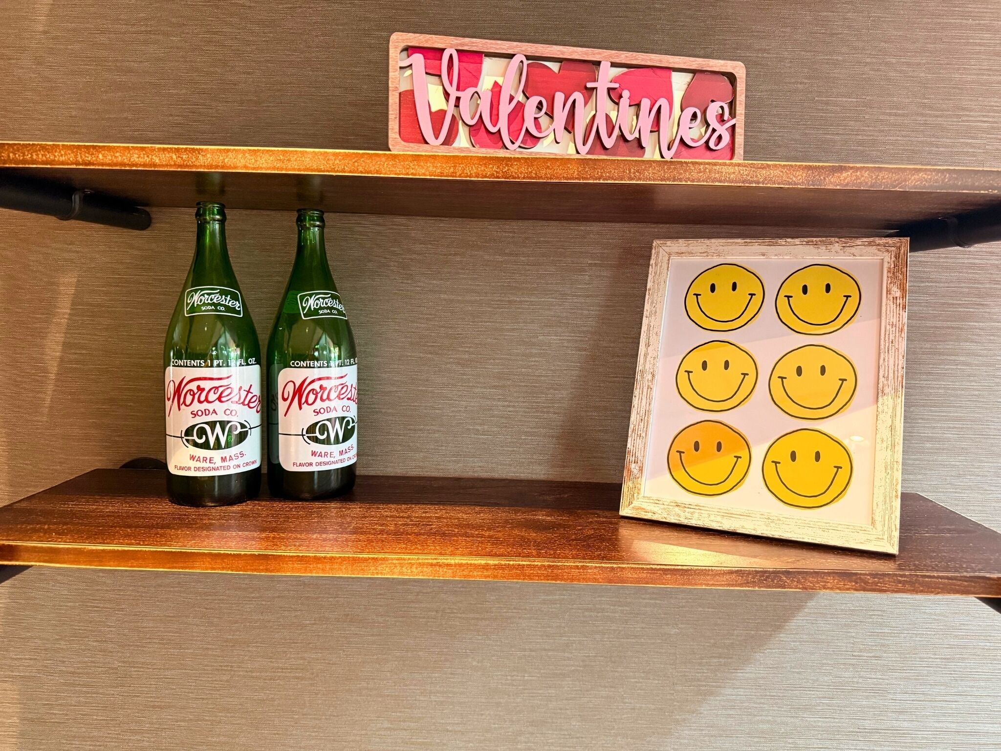 Two bottles of Norcester Soda Co. on a shelf, a 'Valentines' sign above, and a framed picture of smiley faces.
