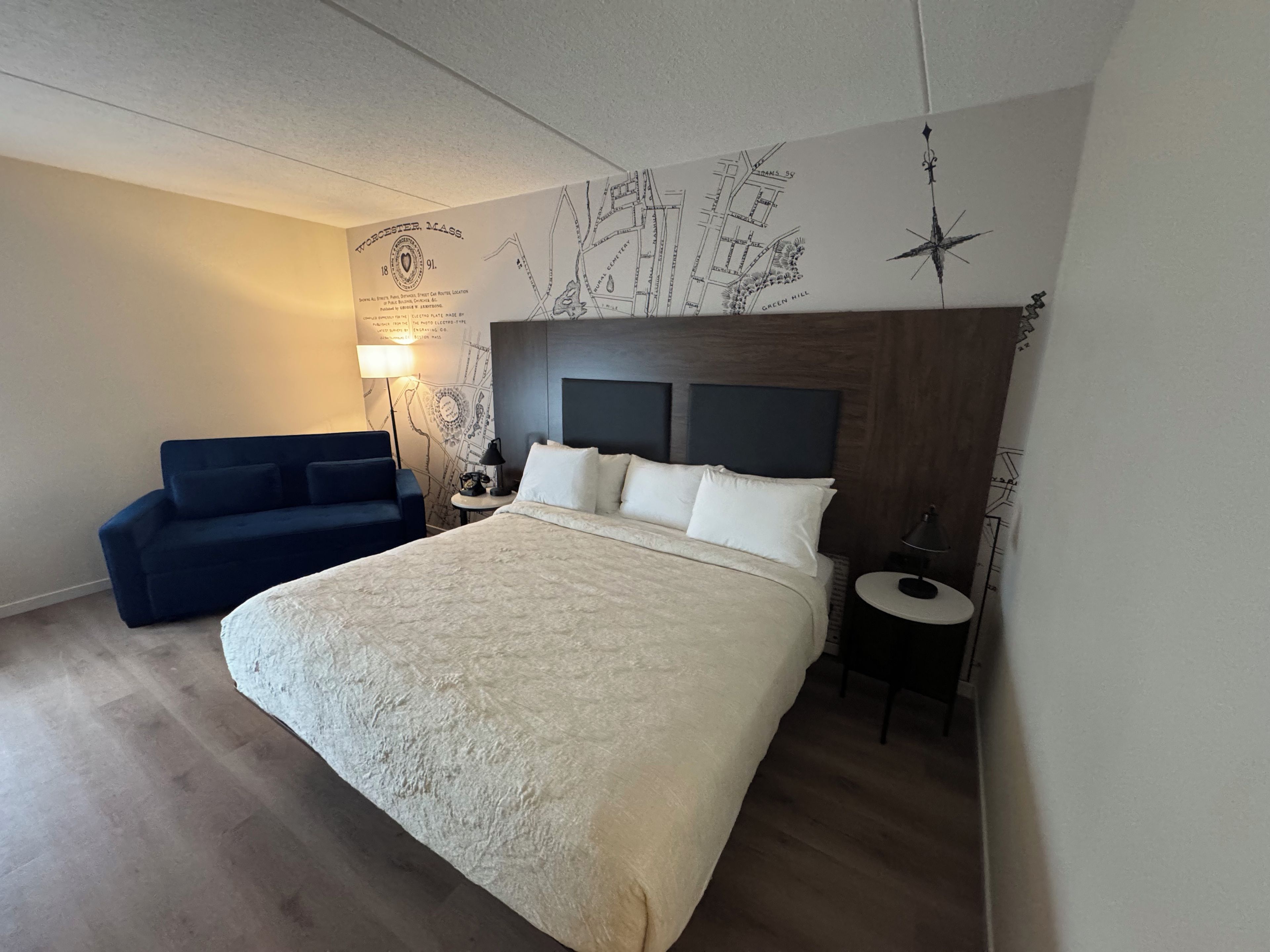Hotellihuone, jossa on suuri sänky, sininen sohva, yöpöytä, lamppu ja seinälle ripustettu kartta.