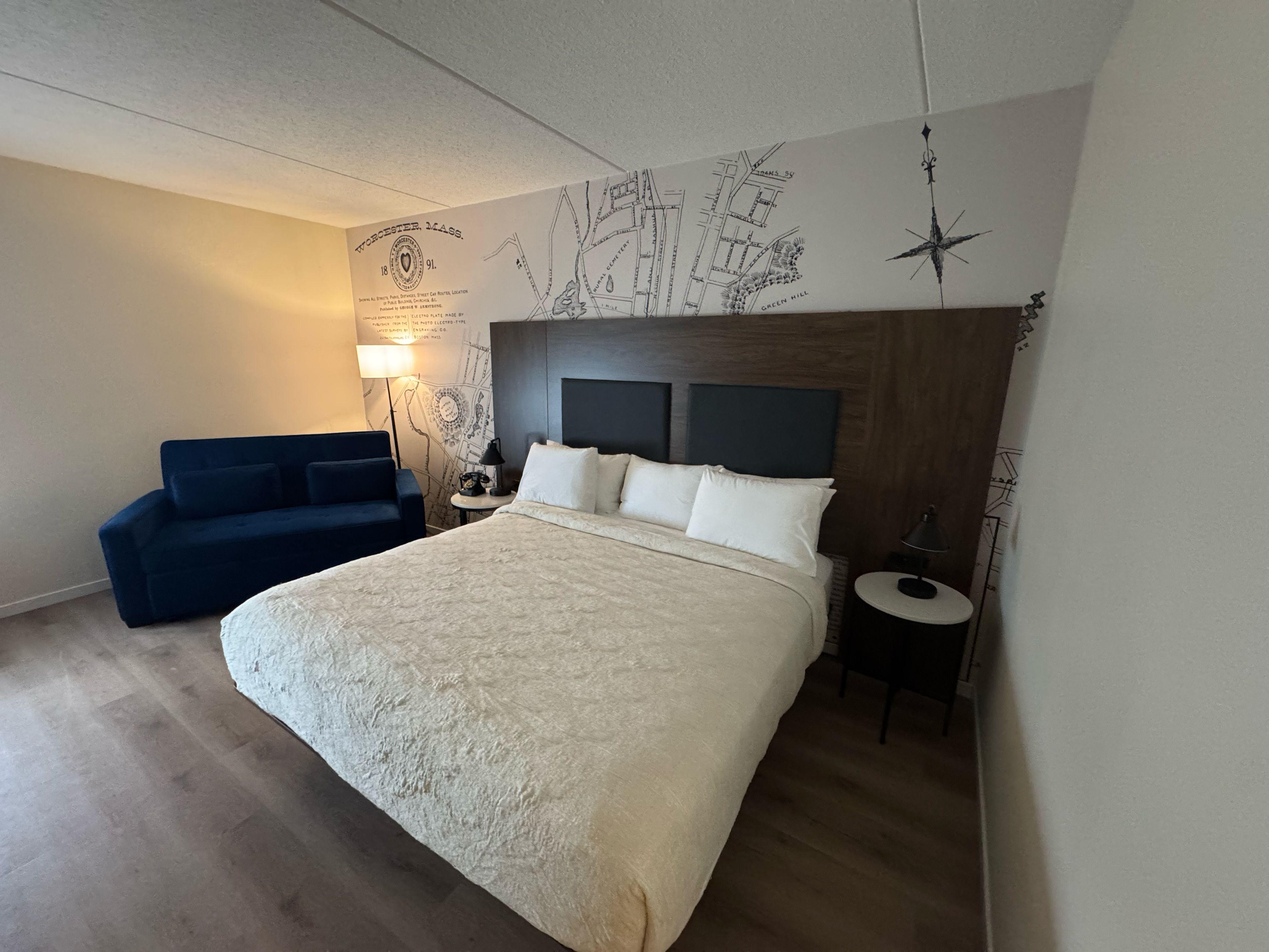 Habitación de hotel con una cama grande, un sofá azul, una mesita de noche y un mapa mural.