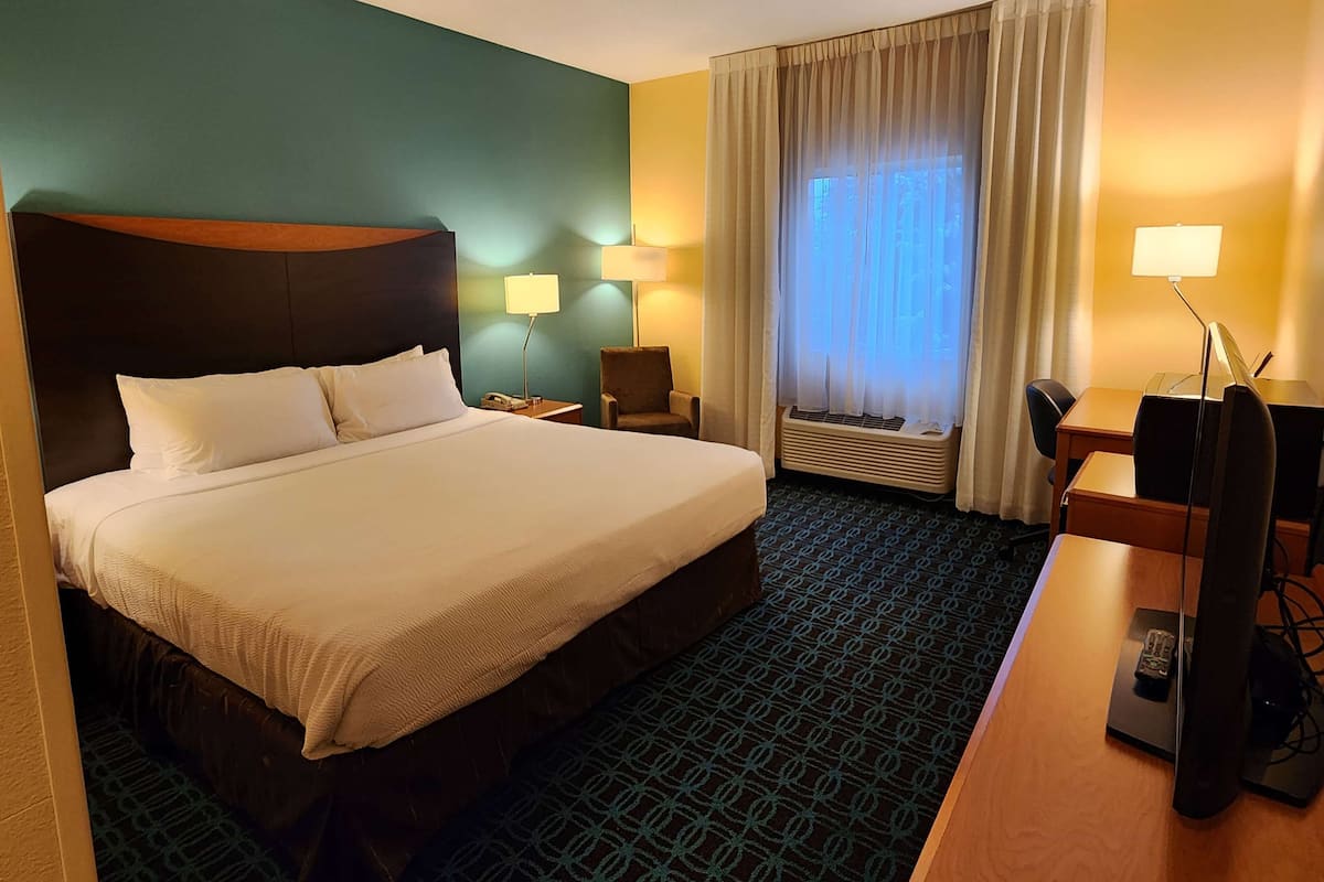 A hotel room with a large bed, a desk with a chair, a lamp, and a window with curtains.