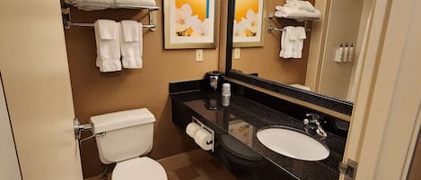 Un baño moderno con inodoro, lavamanos y un espejo grande. Hay toallas en un estante y un cuadro en la pared.