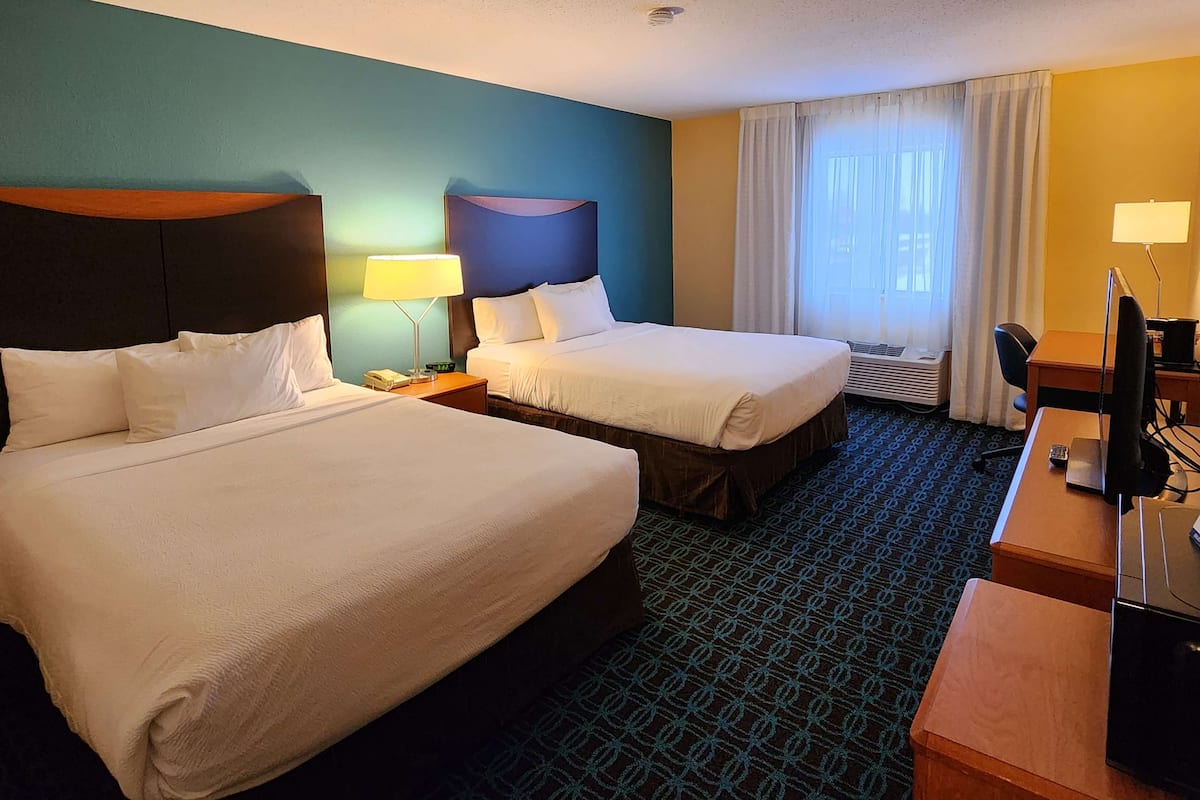 A hotel room with two beds, a desk, a chair, a lamp, and a window with curtains.