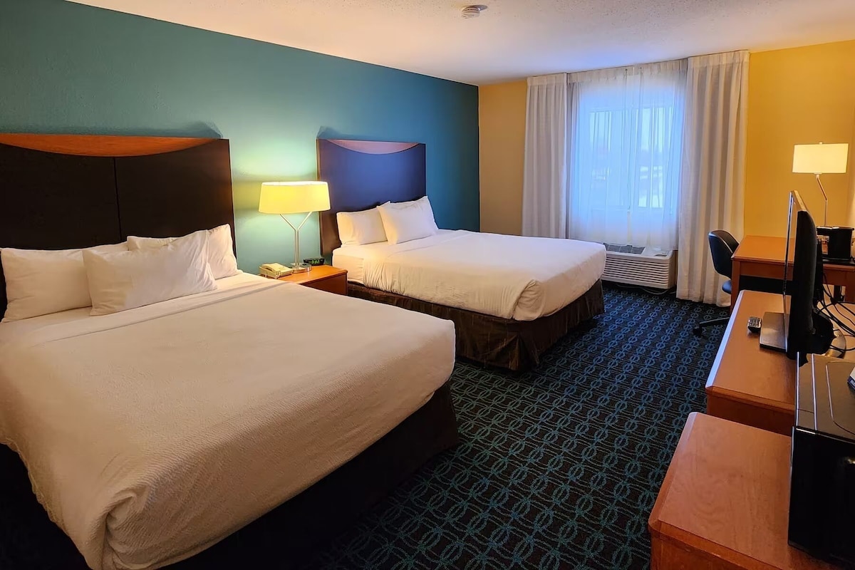 Habitación de hotel con dos camas, un escritorio, una silla, una lámpara y una ventana con cortinas.
