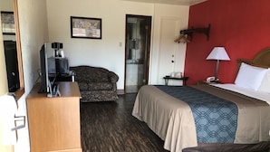 Habitación de hotel con cama, escritorio con computadora, sofá, pared de acento roja y puerta que da a un baño.