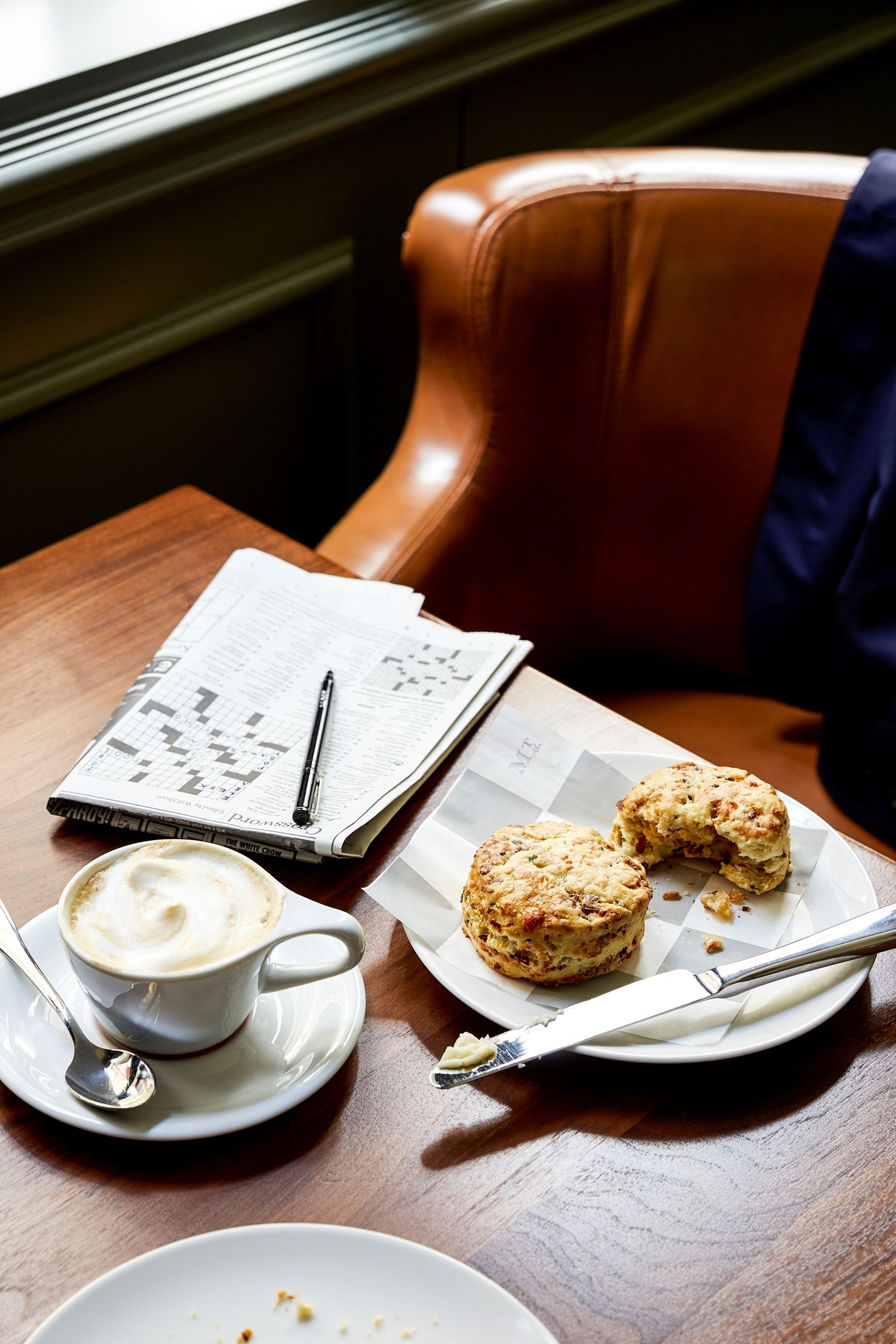 Ein Tisch mit einer Zeitung, einer Tasse Kaffee, zwei Scones und einem Teller mit Messer und Löffel.