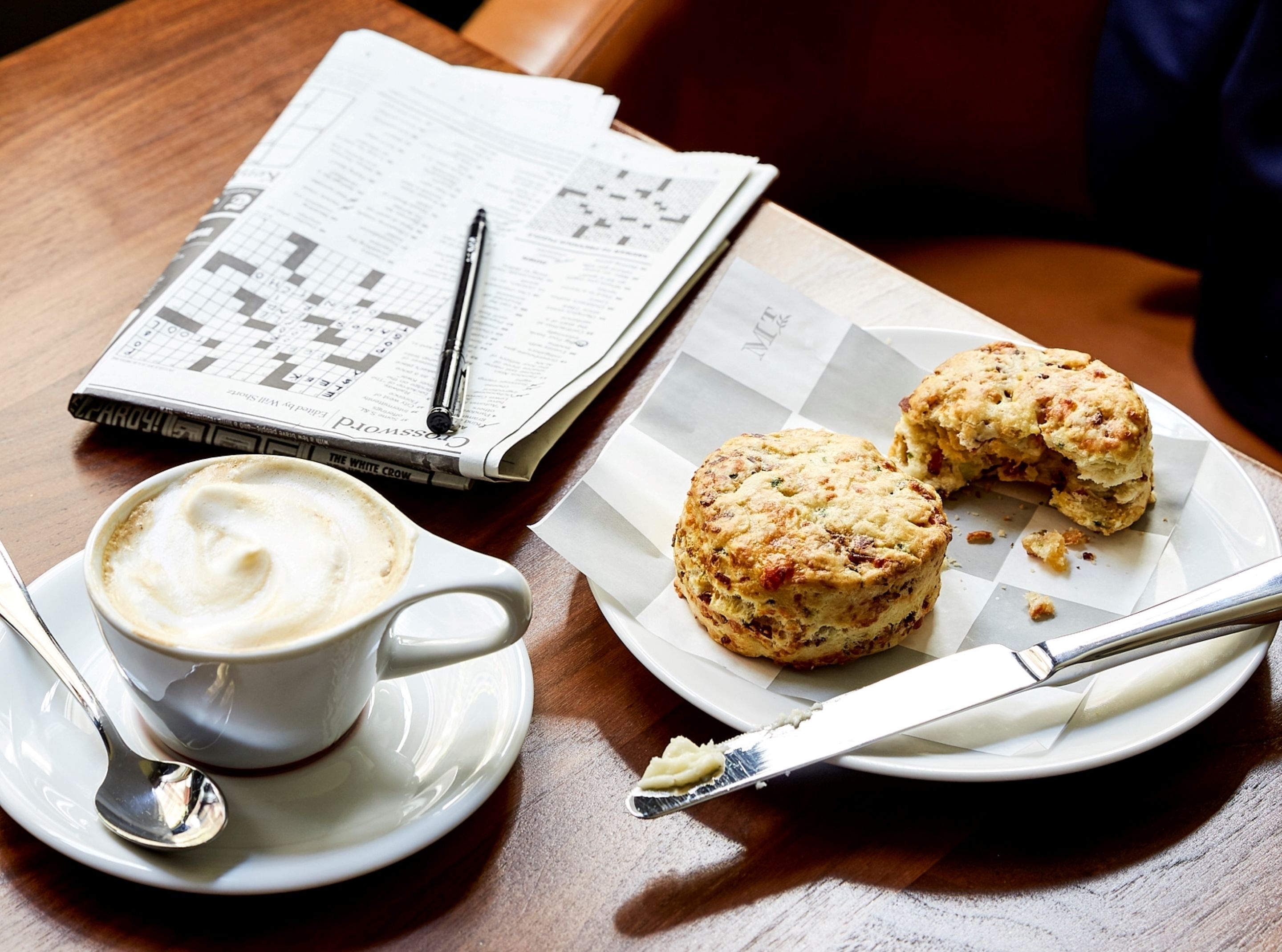 A table with a cup of coffee, a plate of scones, a newspaper, and a pen.