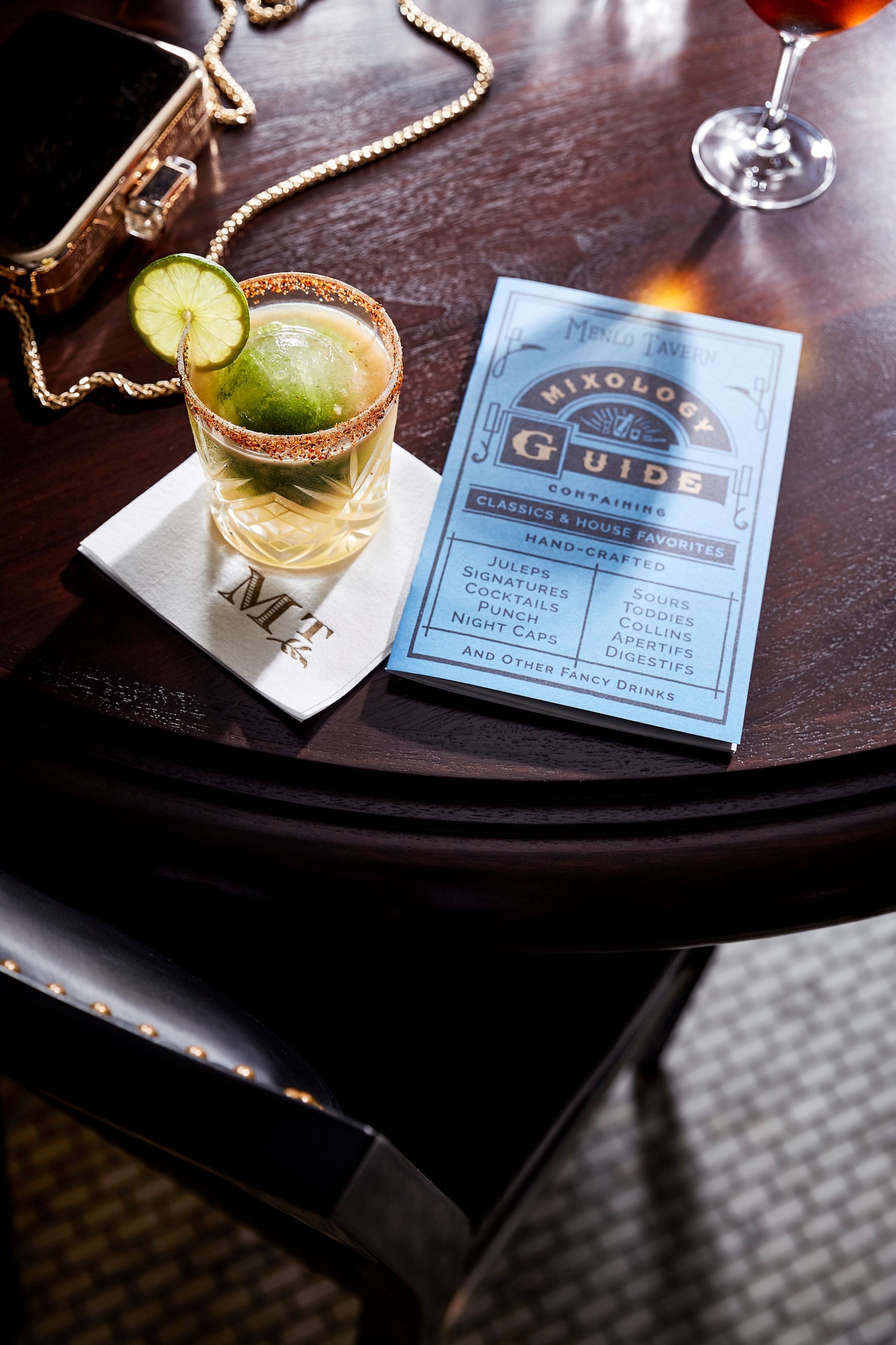 A cocktail glass with a lime garnish on a napkin, a menu titled "Mixology Guide," and a gold chain purse on a wooden table.