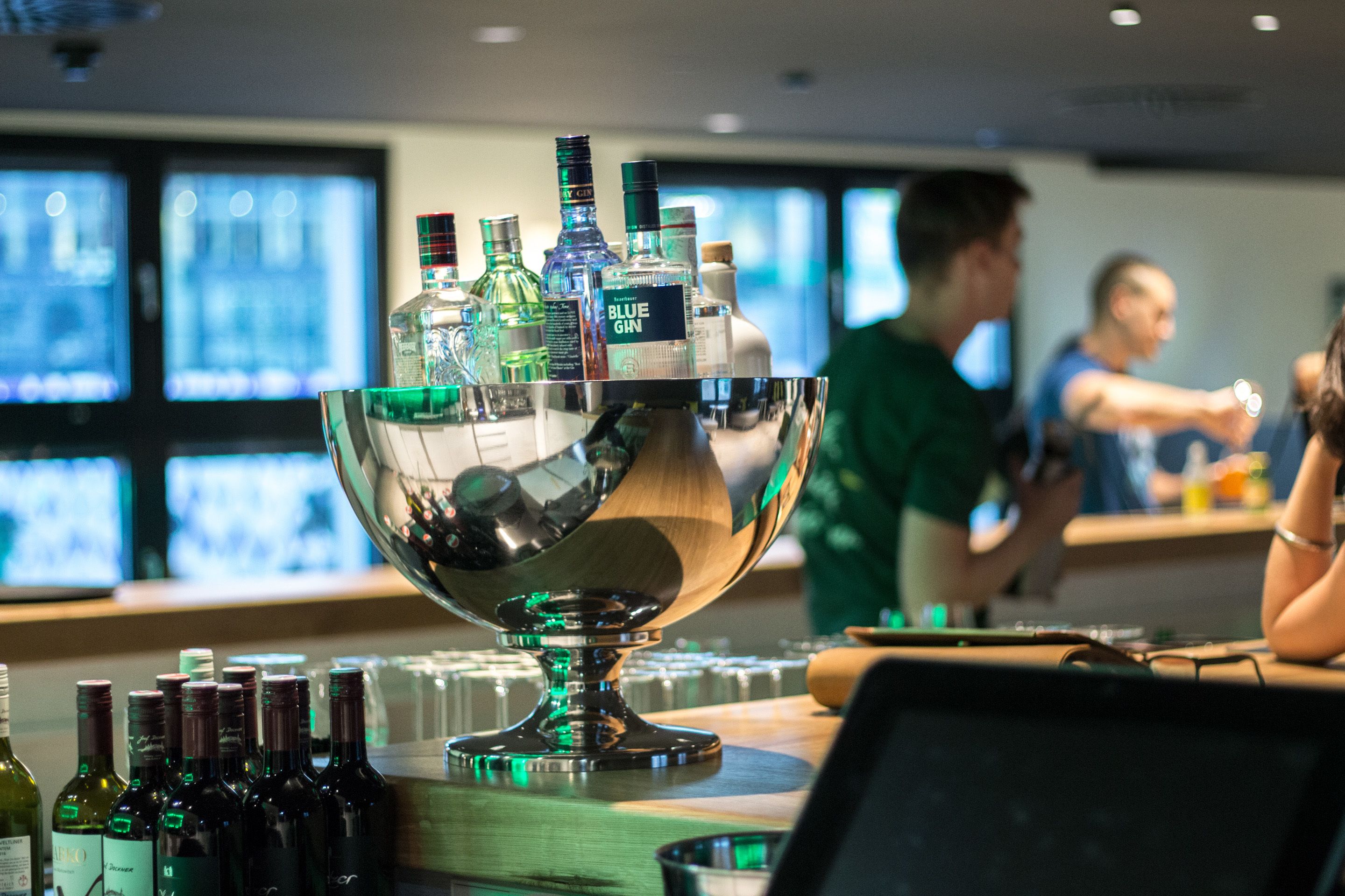 Un área de bar con un recipiente metálico grande lleno de varias botellas, presumiblemente con bebidas. Al fondo, hay varias pantallas mostrando contenido.