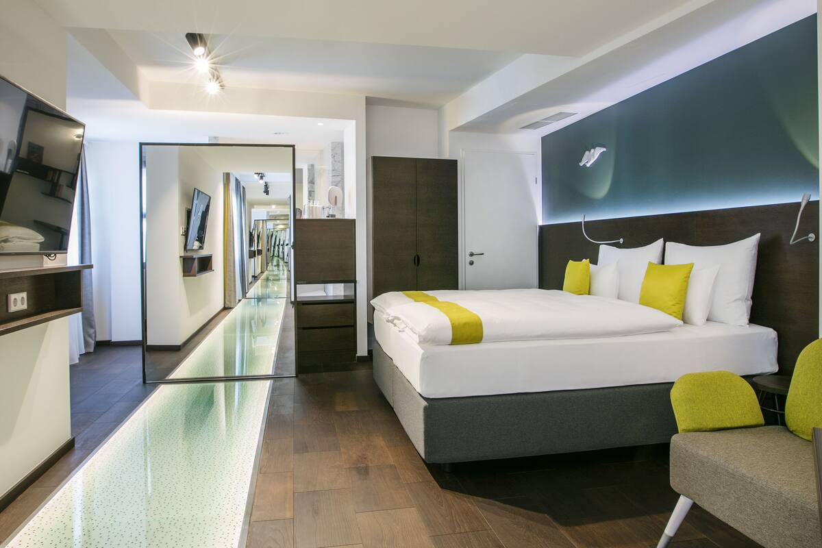 A modern hotel room with a large bed, a sofa, a mirror, and a long hallway leading to a bathroom.