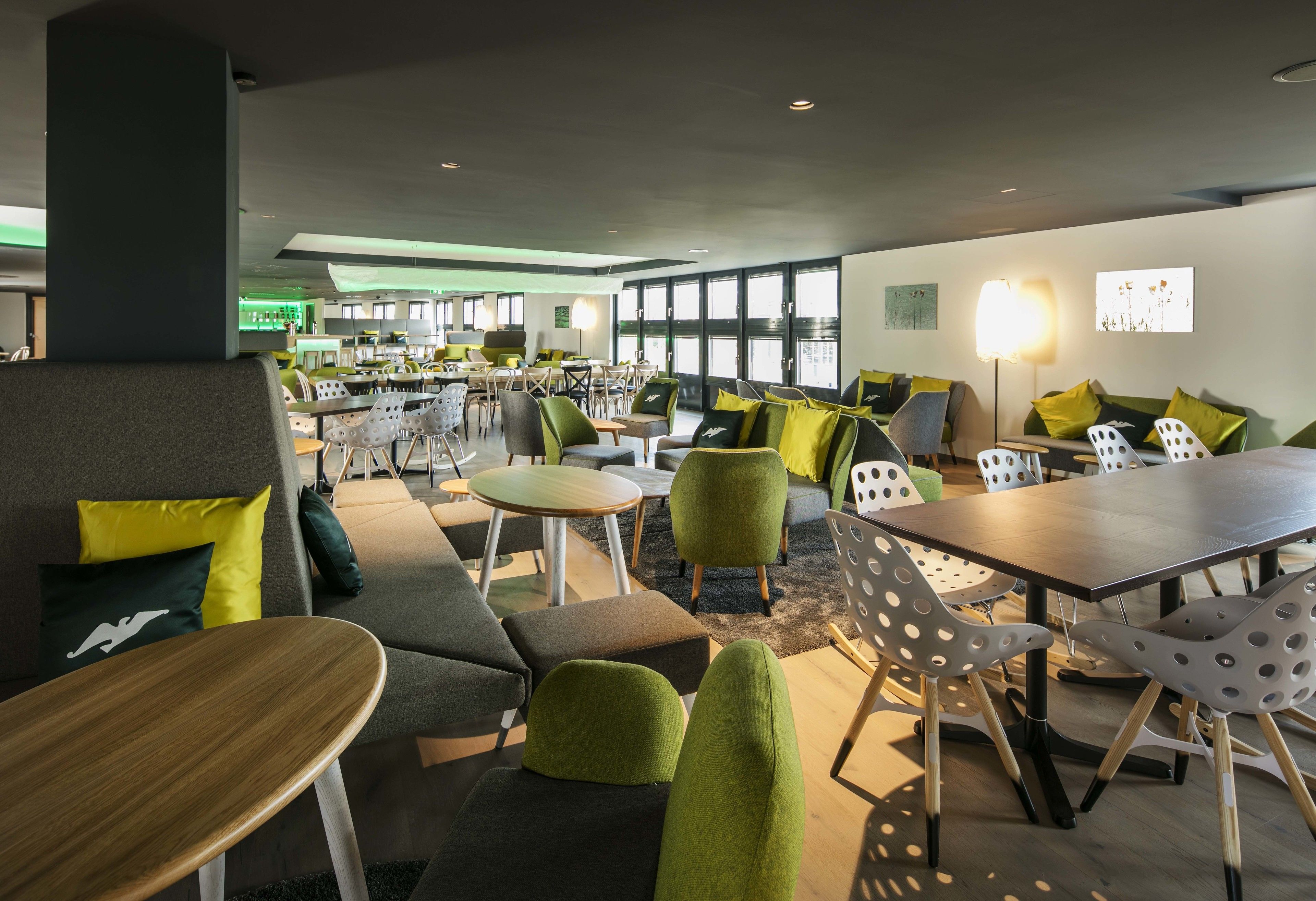 Un espace de détente moderne avec des sièges verts et jaunes, des tables en bois et une grande fenêtre laissant entrer la lumière naturelle.
