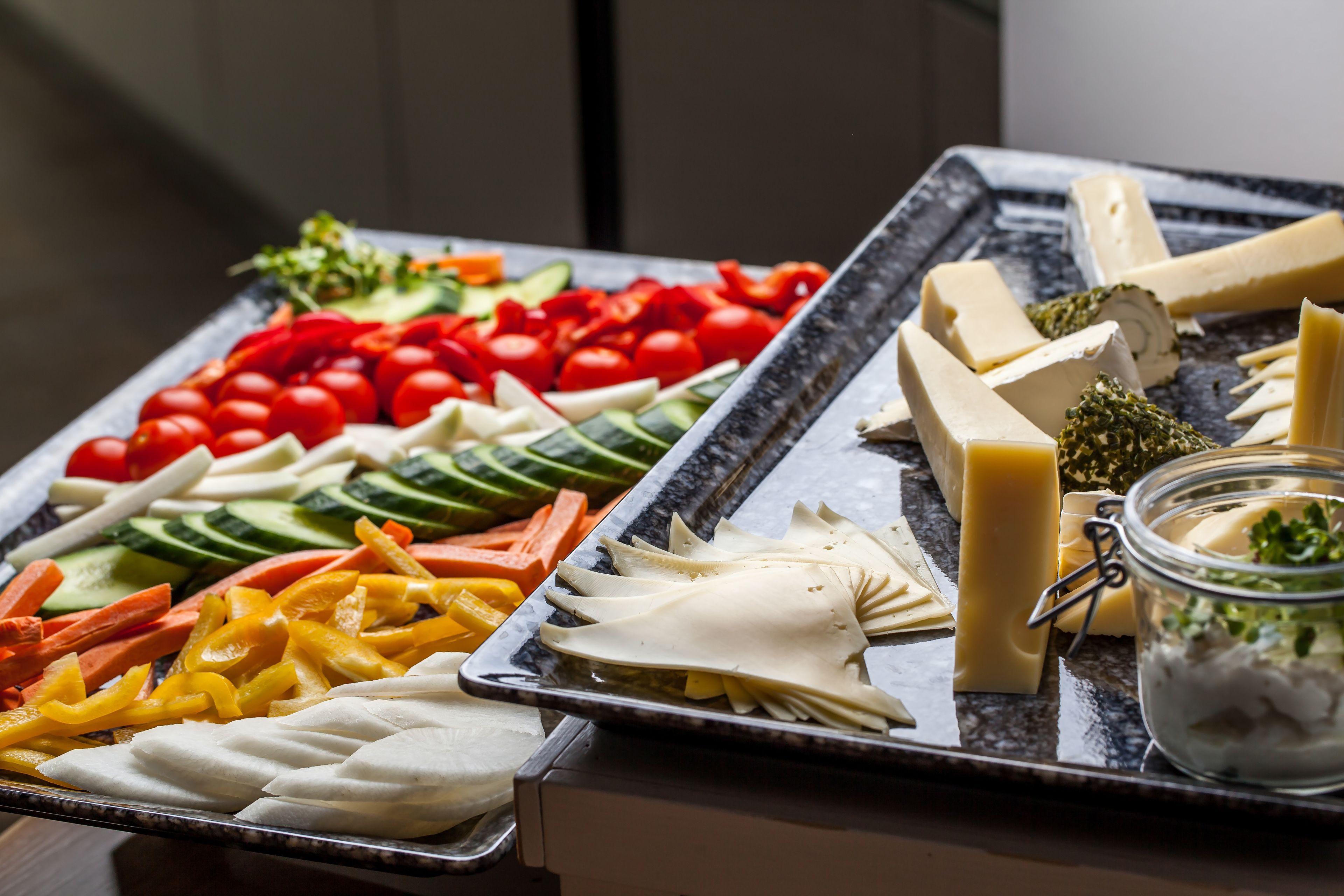 A buffet with various cheeses, vegetables, and a jar of olives.