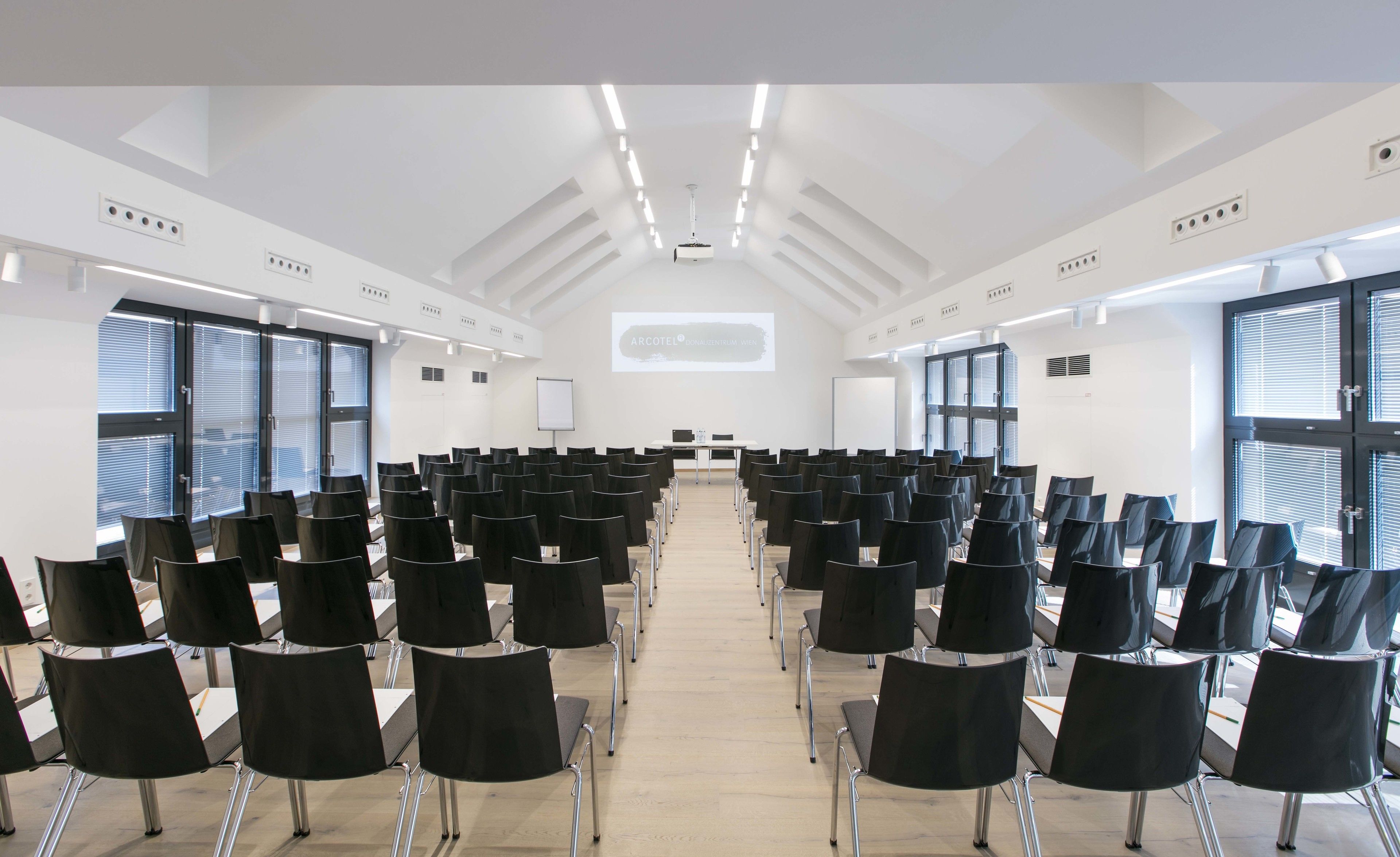 A conference room with rows of black chairs, a white projection screen, and large windows with blinds.