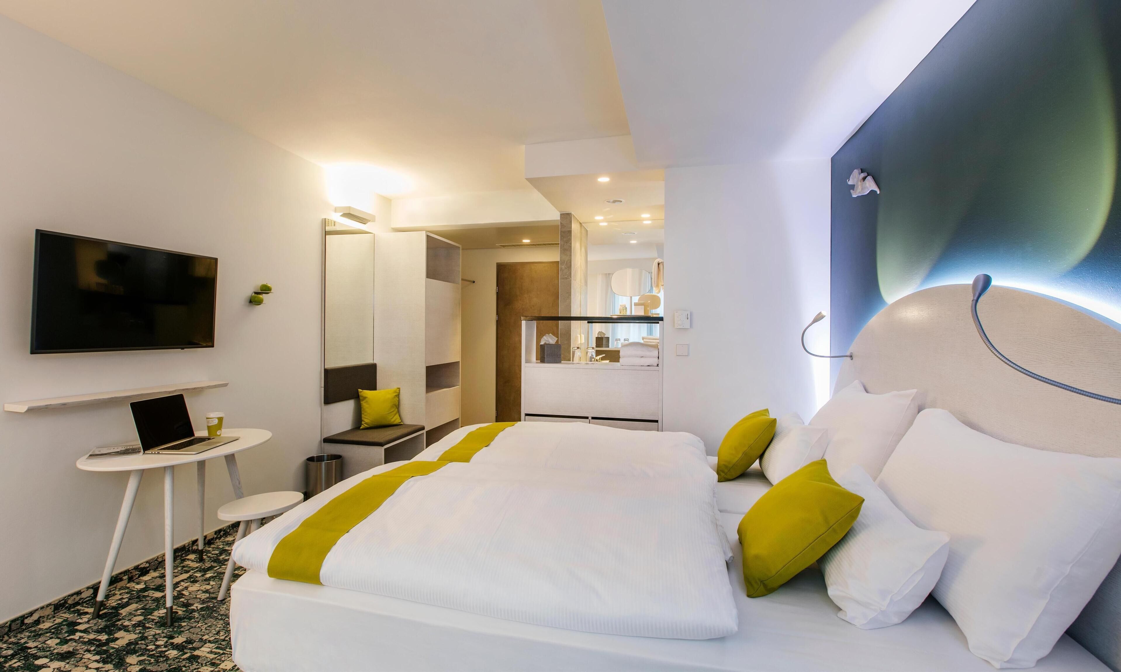 Une chambre d’hôtel moderne dotée d’un grand lit, d’un bureau avec un ordinateur portable, d’une télévision à écran plat et d’un miroir dans la salle de bain.