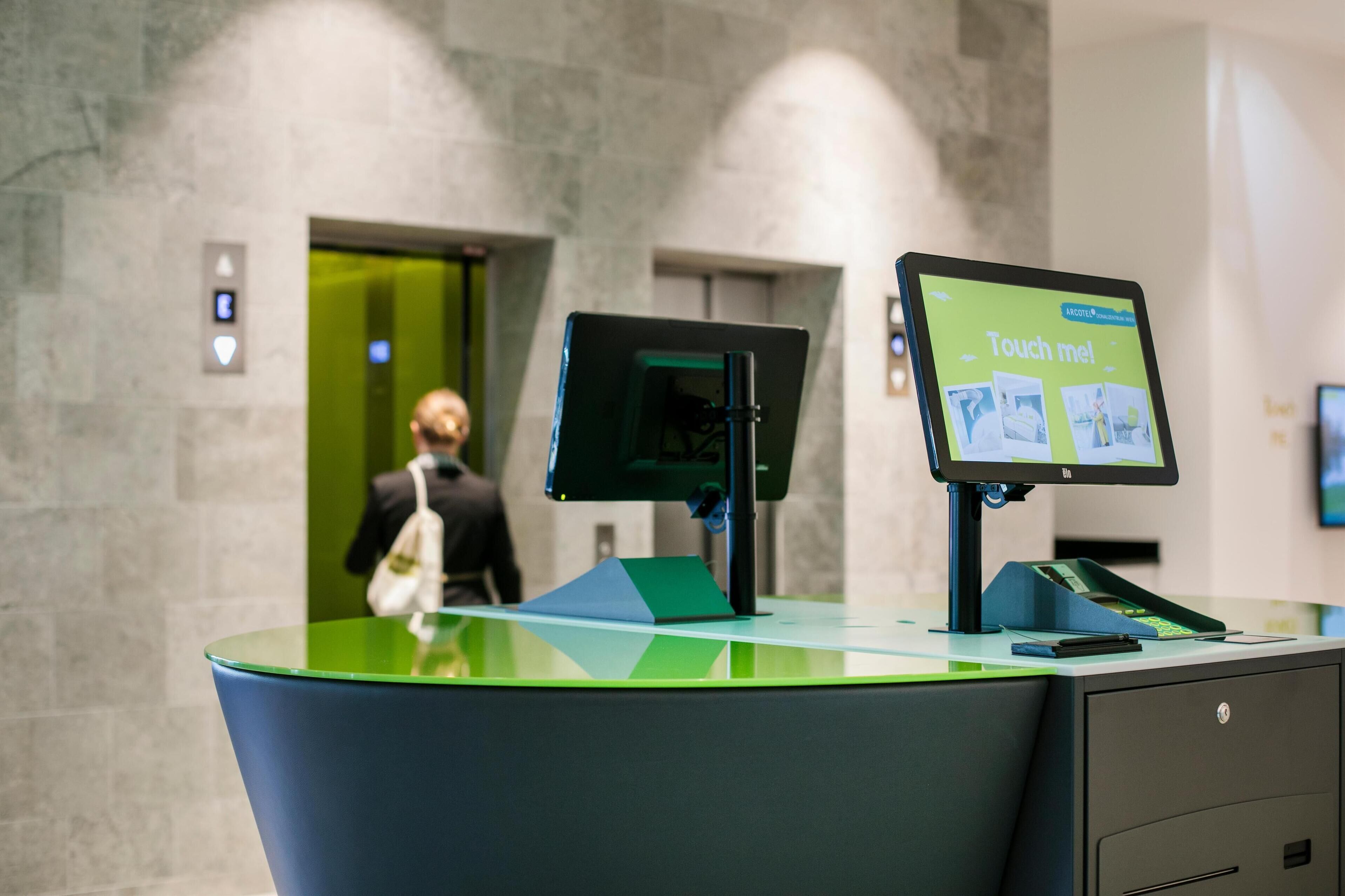 A modern reception desk with a digital touchscreen display, a green and black color scheme, and a marble wall in the background.
