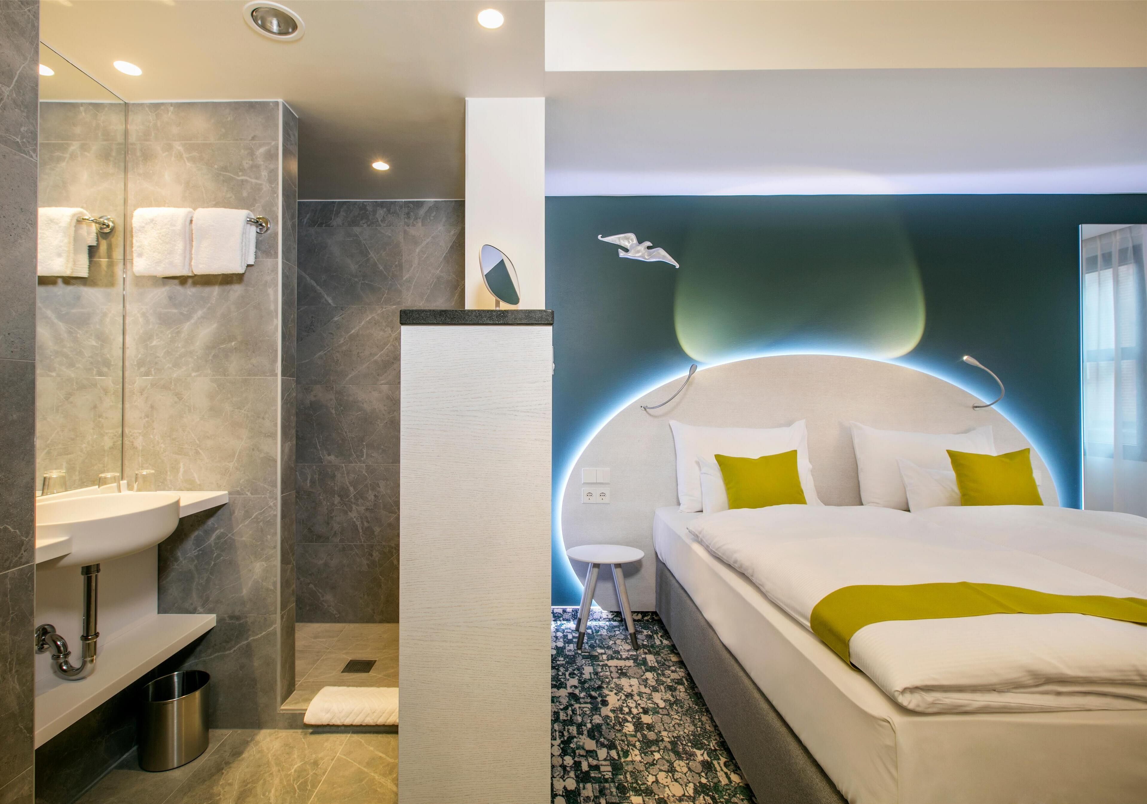 A modern hotel room with a large bed, a separate bathroom with a sink, and a small stool.