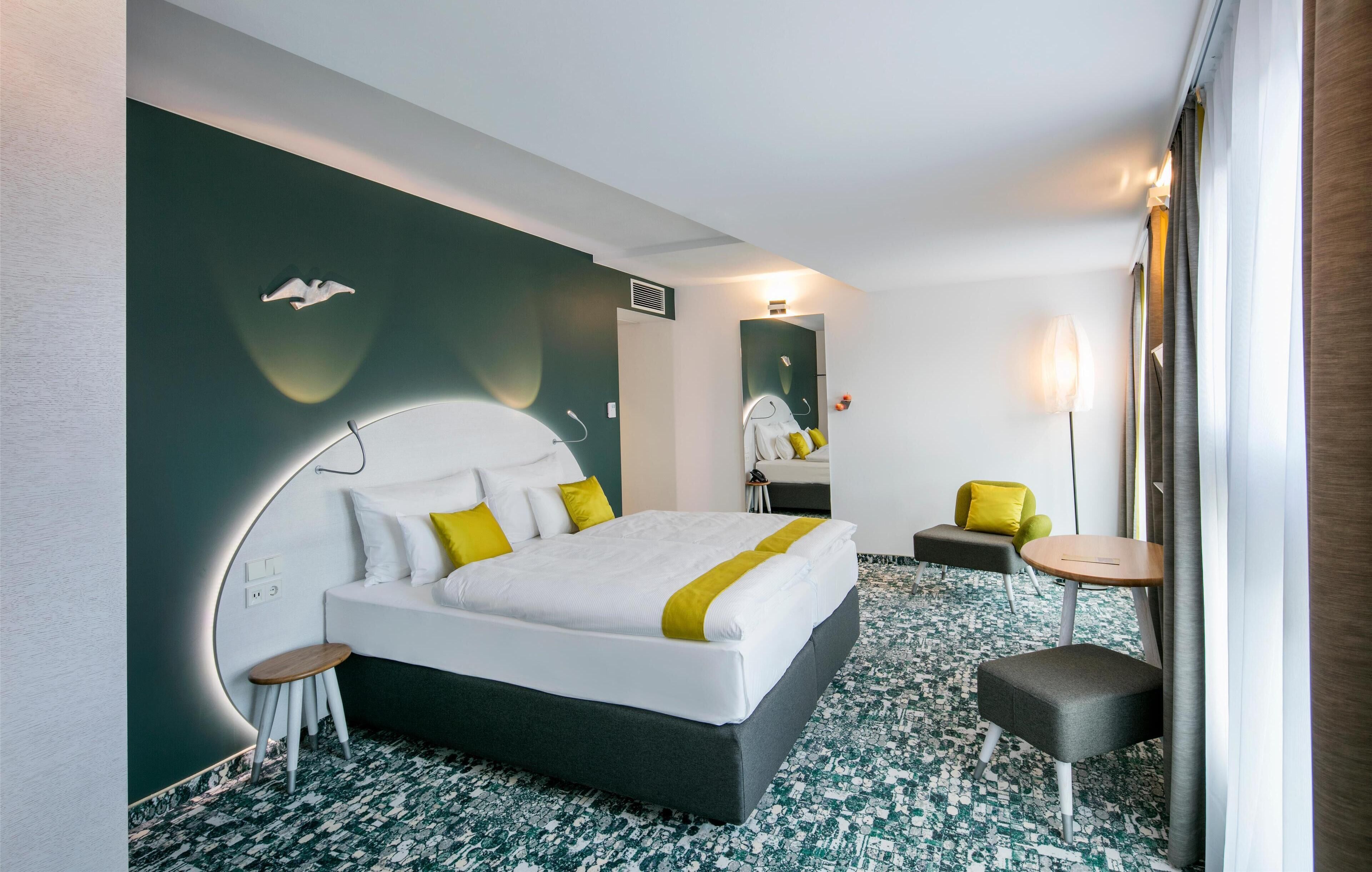 Ein modernes Hotelzimmer mit einem großen Bett, einem Nachttisch, einem Sessel, einem kleinen Hocker, einem Spiegel und einer Lampe.