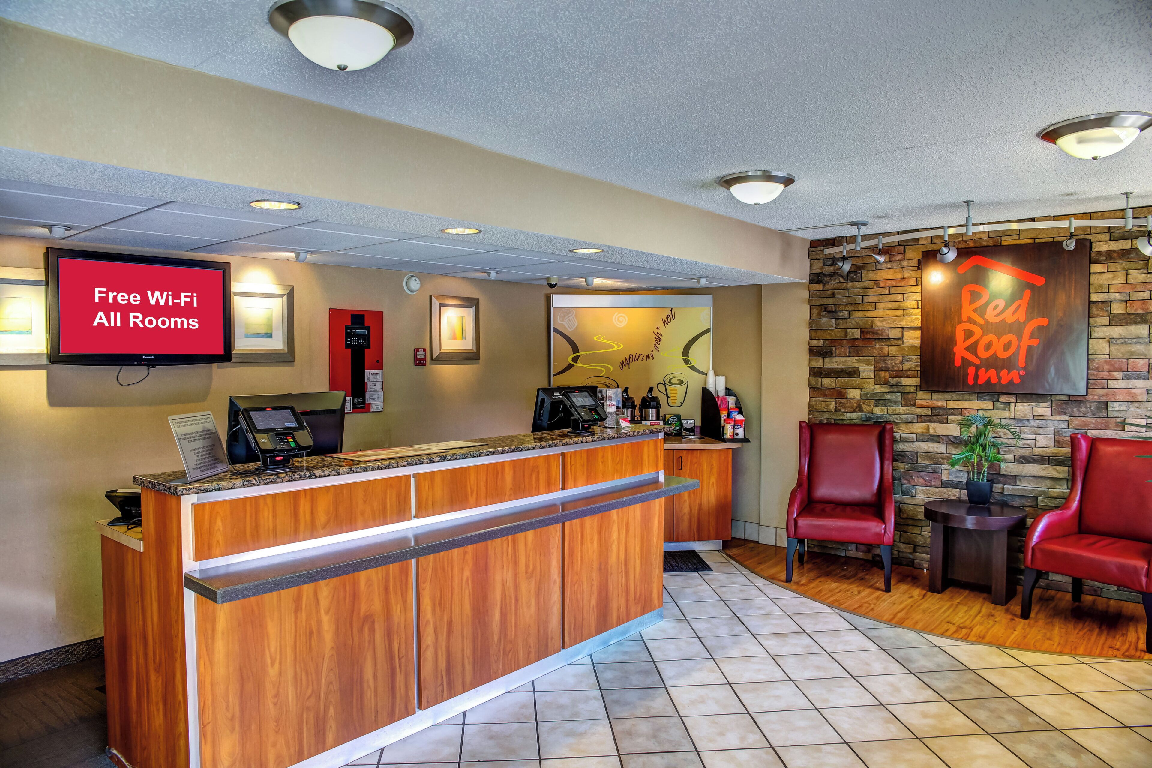 Uma área de recepção de hotel com uma tela digital exibindo 'Wi-Fi Gratuito em Todos os Quartos', um balcão de atendimento, um alarme de incêndio e a placa da Red Roof Inn.