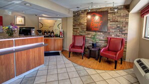 A reception area with a wooden counter, two red chairs, a small table with a plant, and a wall-mounted sign. - Red Roof Inn Pensacola – I-10 at Davis Highway (Pensacola)