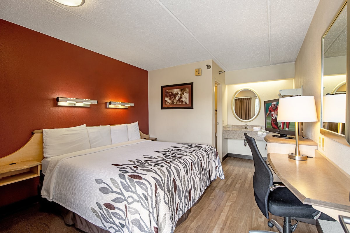 Una habitación de hotel con cama, escritorio con silla, espejo y televisor.