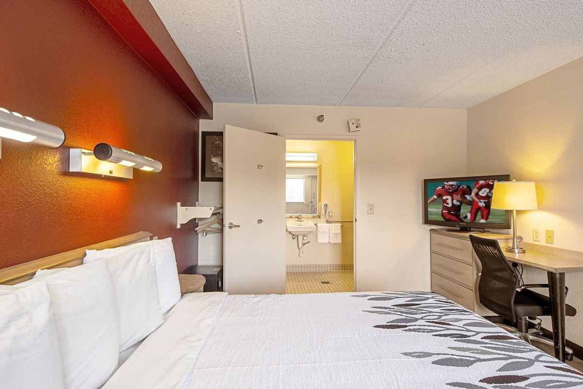 Habitación de hotel con una cama, un escritorio con silla, televisi ón y baño visible a través de una puerta abierta.