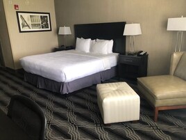Una habitación de hotel con una cama grande, dos mesitas de noche con lámparas, una silla y un pequeño reposapiés.