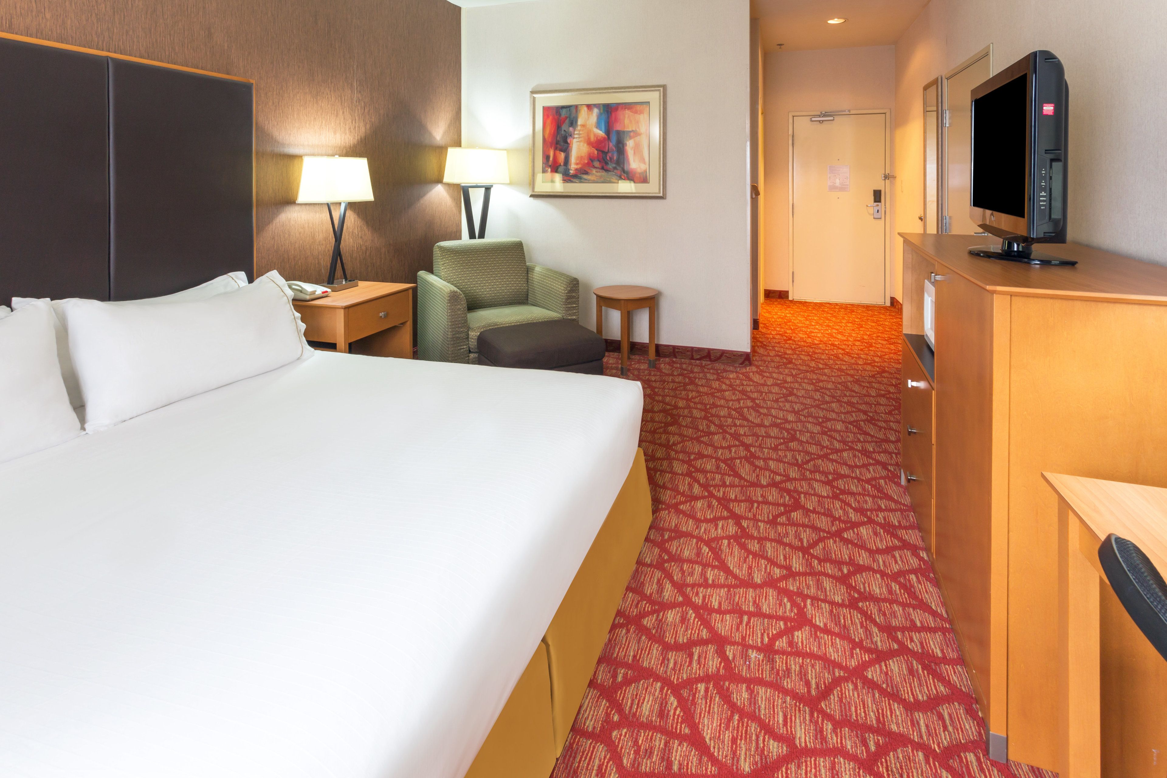 A hotel room with a large bed, a desk with a lamp, a television, and a small chair.