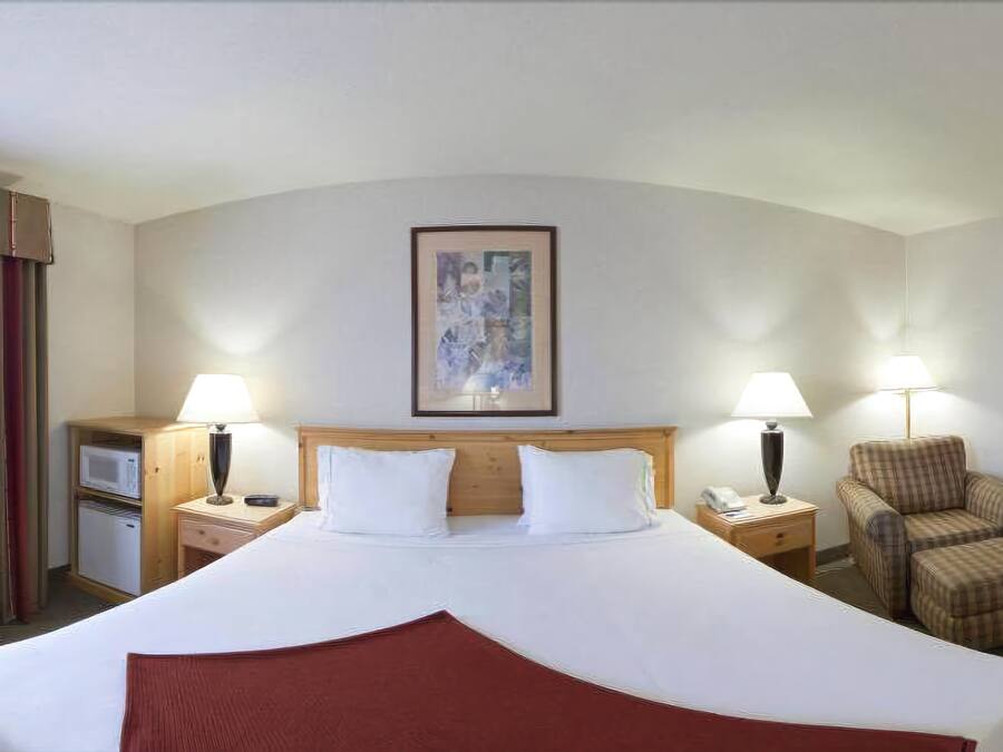 Una habitació d'hotel amb un llit gran, dues taules de nit amb làmpades, una cadira, una taula petita i un quadre amb marc a la paret.