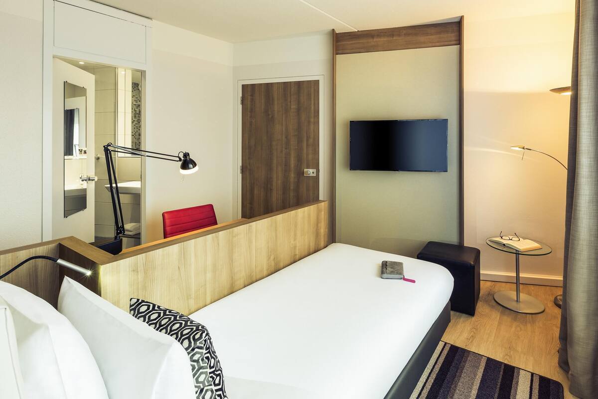 Ein Hotelzimmer mit Bett, Nachttisch, Lampe, Fernseher und Spiegel.