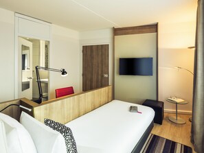 Quarto de hotel com cama, mesa de cabeceira, luminária, televisão e espelho.