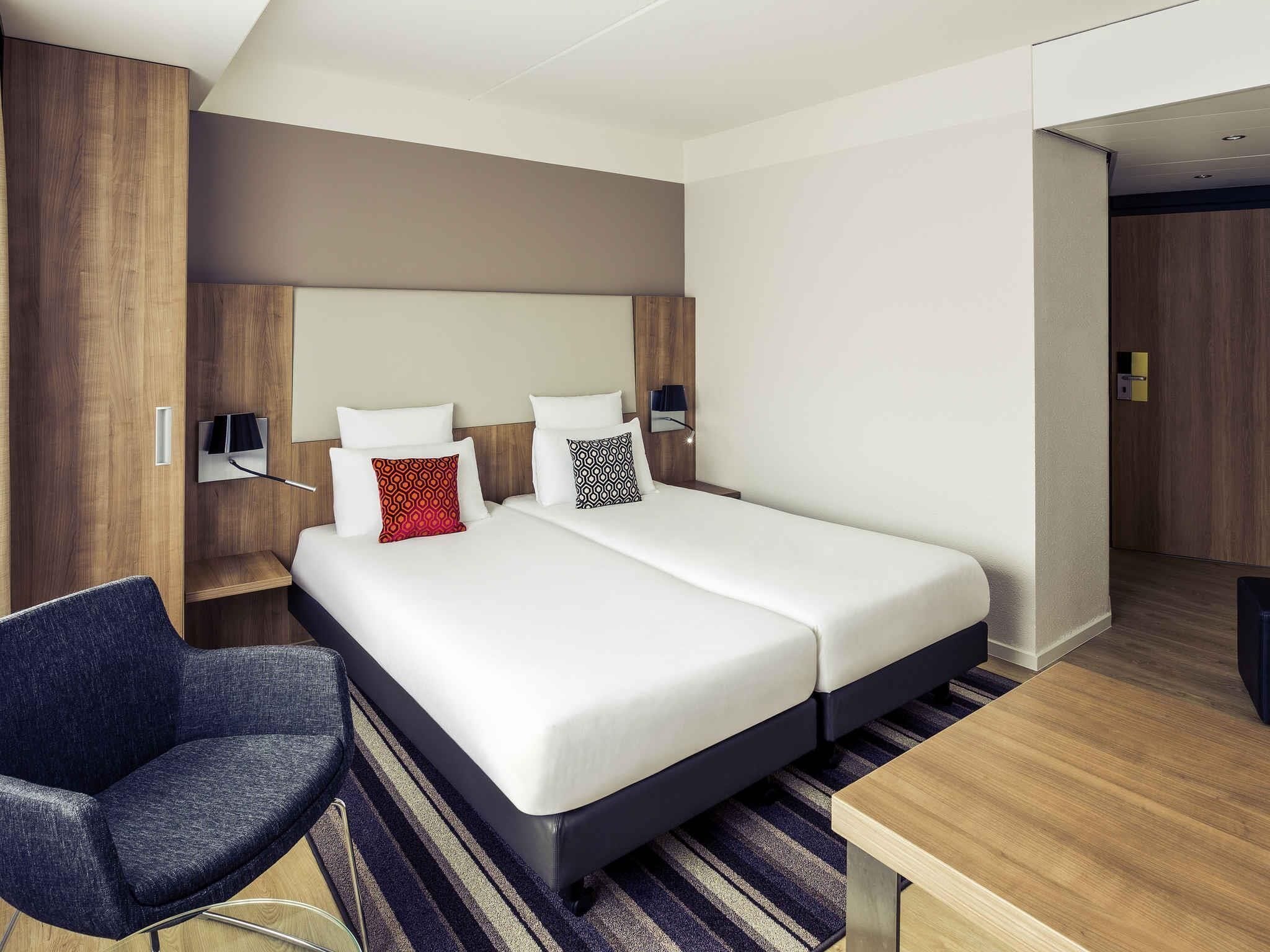 A modern hotel room with a large bed, a desk, a chair, and a wardrobe.